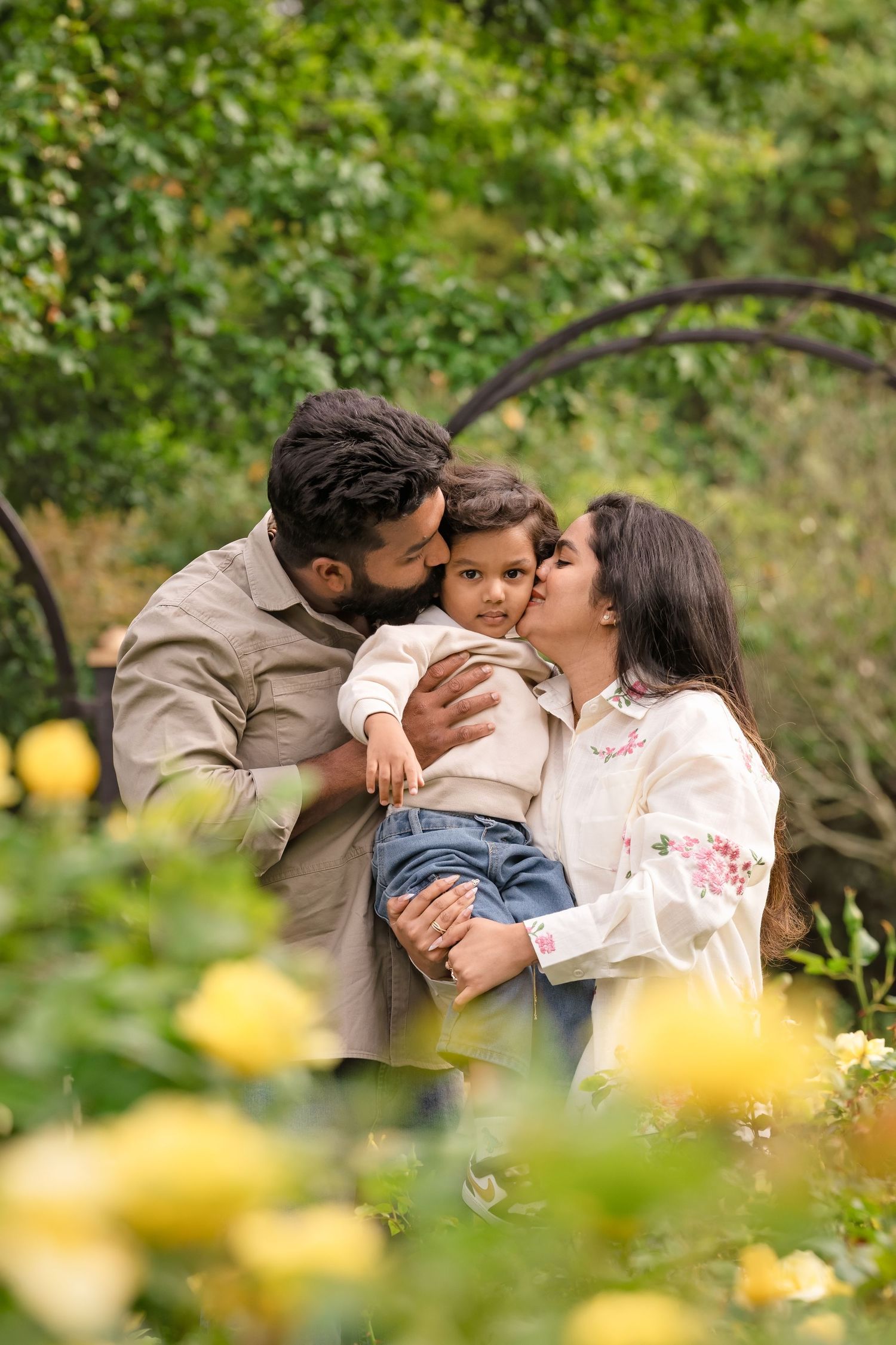 A loving family of three shares kisses outdoors, surrounded by yellow roses and lush green foliage.