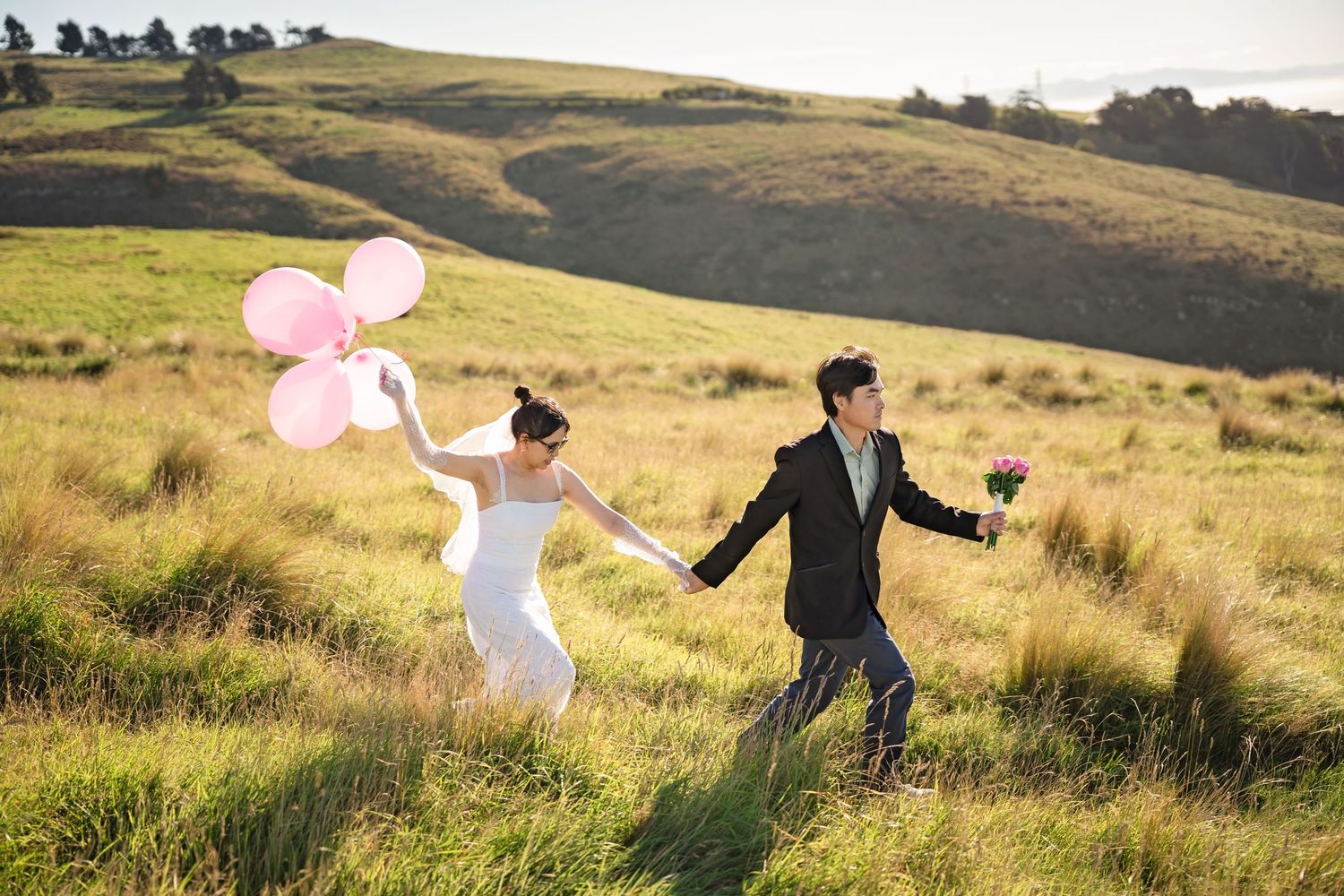 A bride and groom hold hands running through green hills, she carries pink balloons while he holds a bouquet.