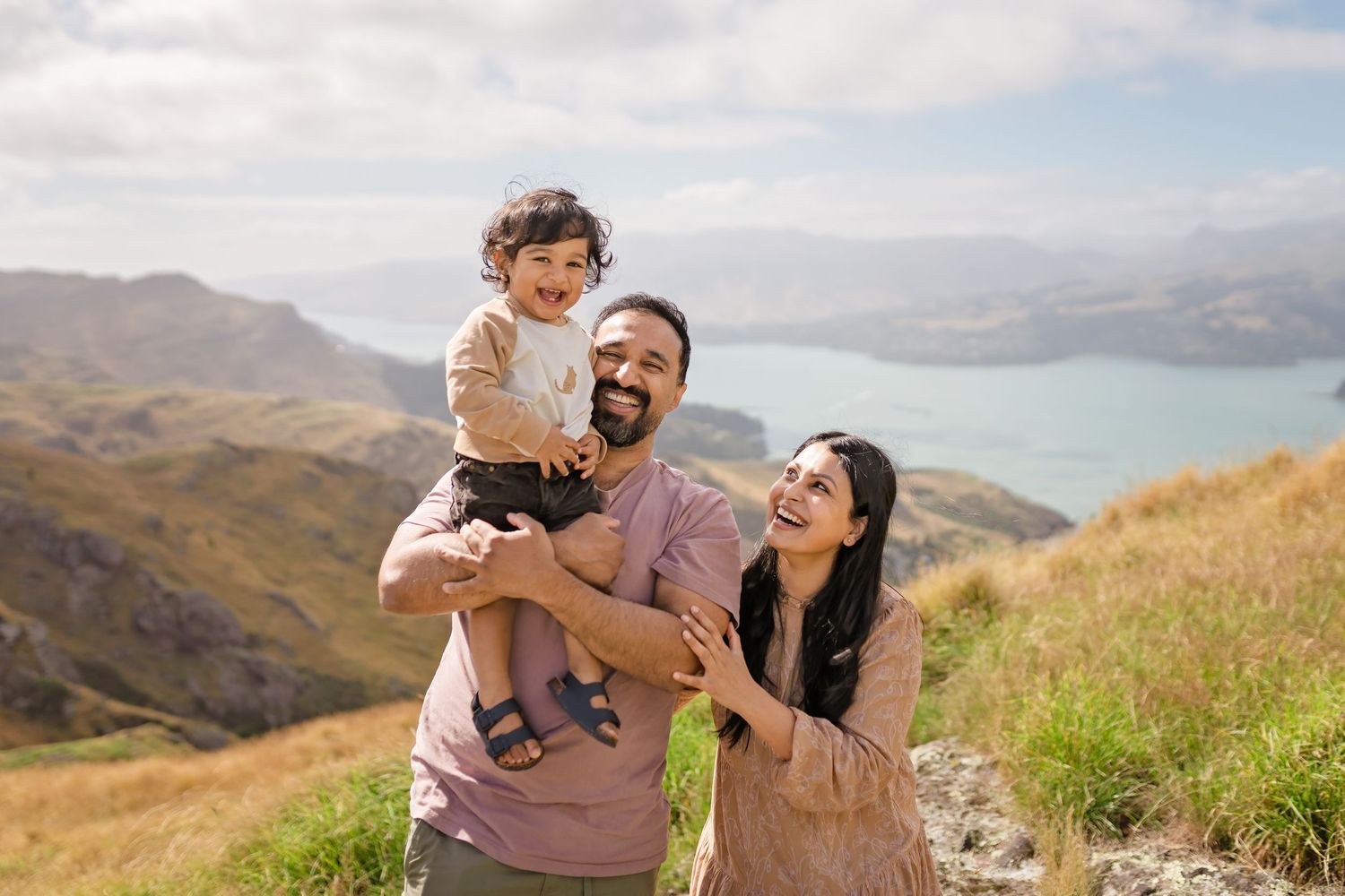 Happy family with a laughing toddler posing on a scenic mountain hillside with a lake and blue sky backdrop.