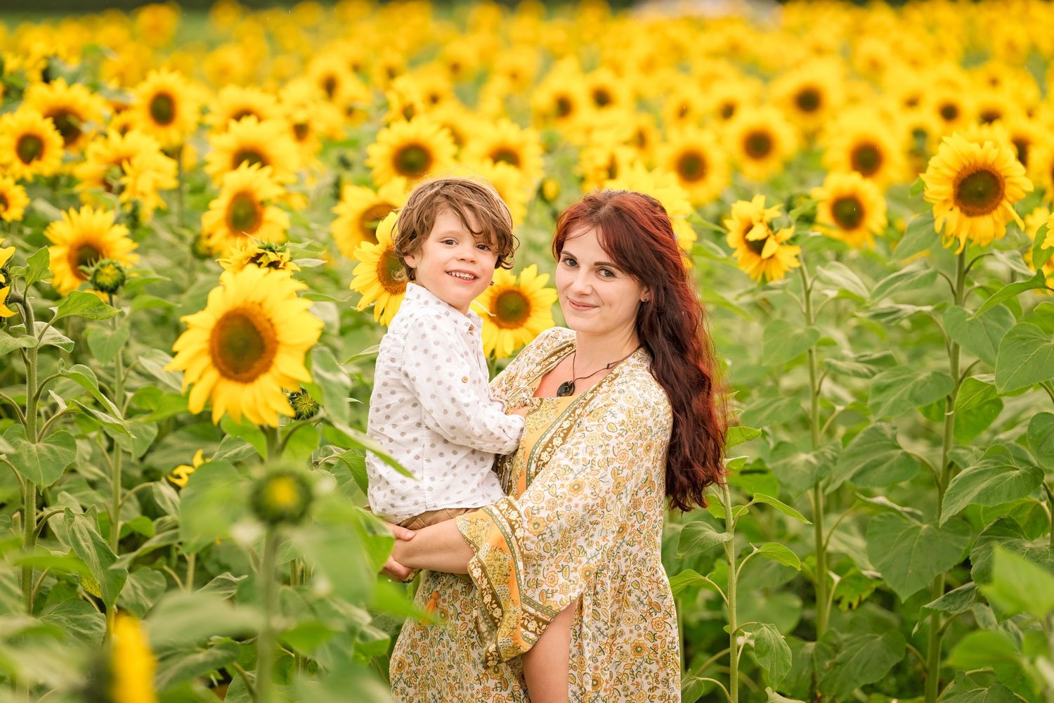 Mother holding child in sunflower field, both wearing floral dresses, smiling outdoors.