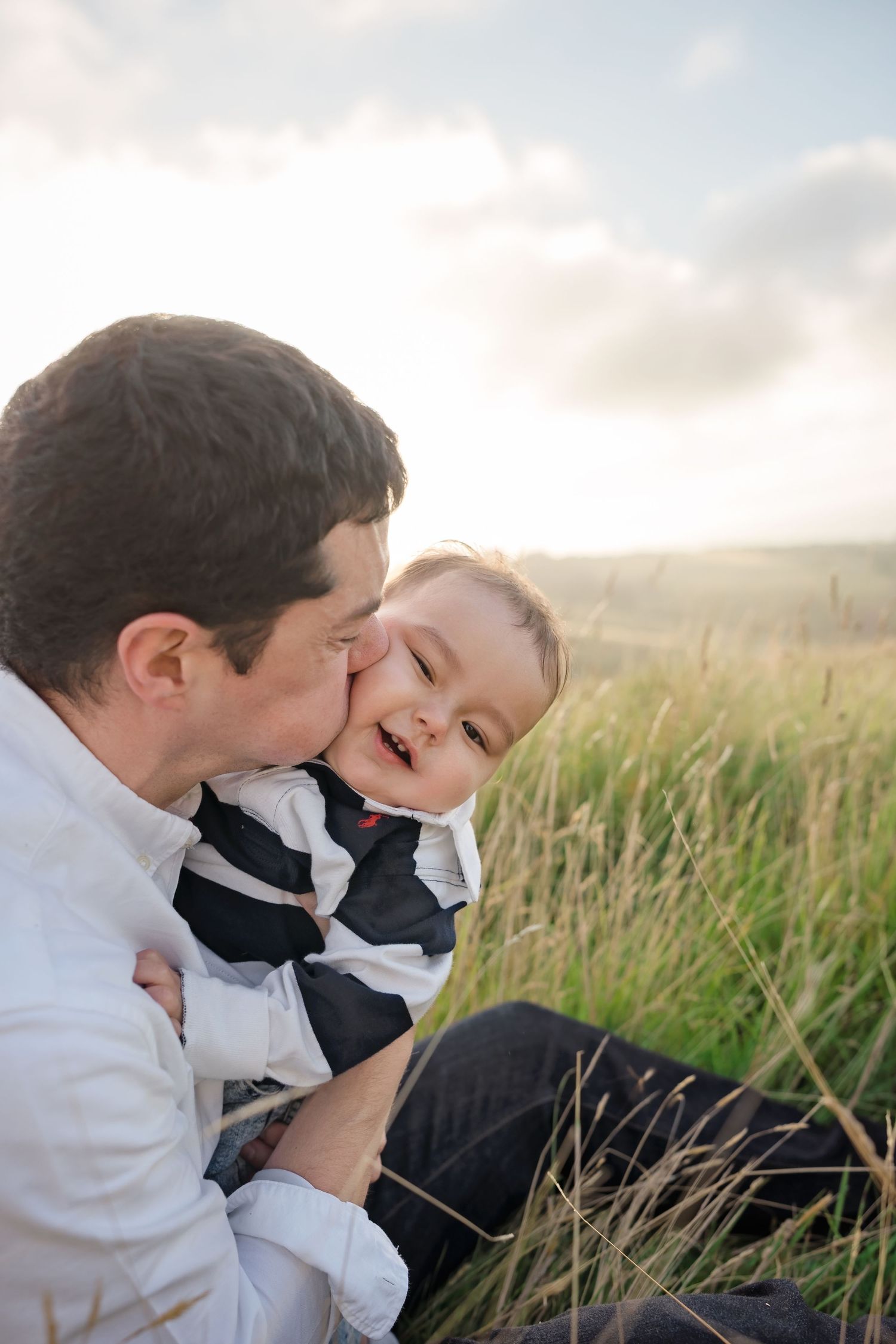 A father kisses his laughing baby in a sunlit grassy field, sharing a joyful outdoor moment.