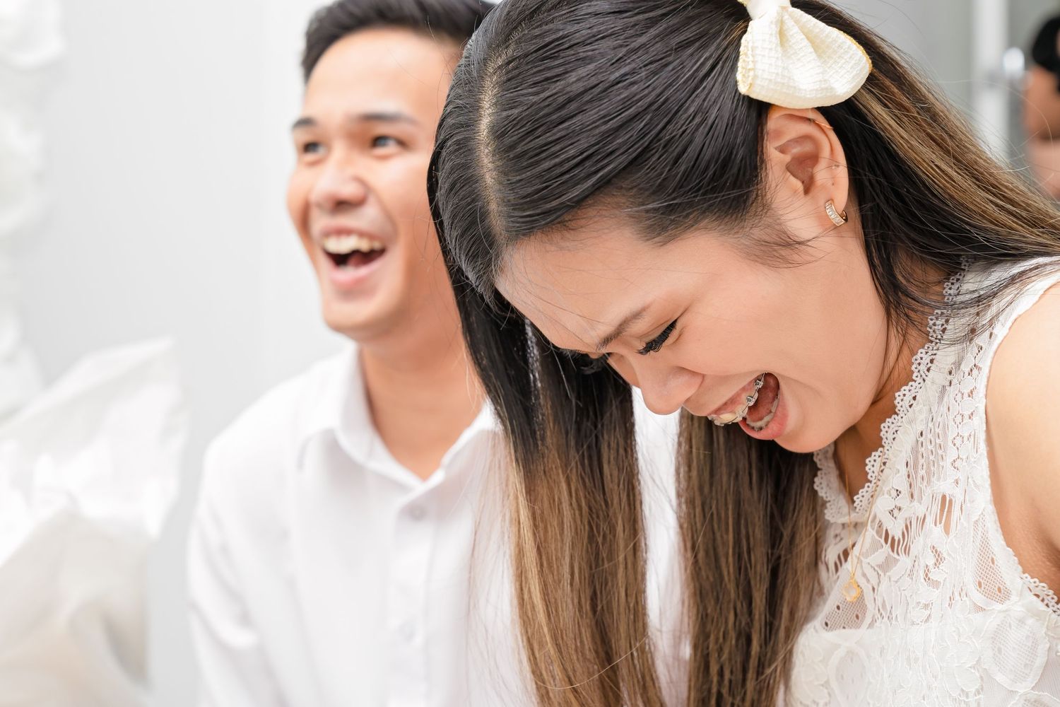 A joyful Asian couple laughing together, dressed in white outfits, sharing a happy moment.