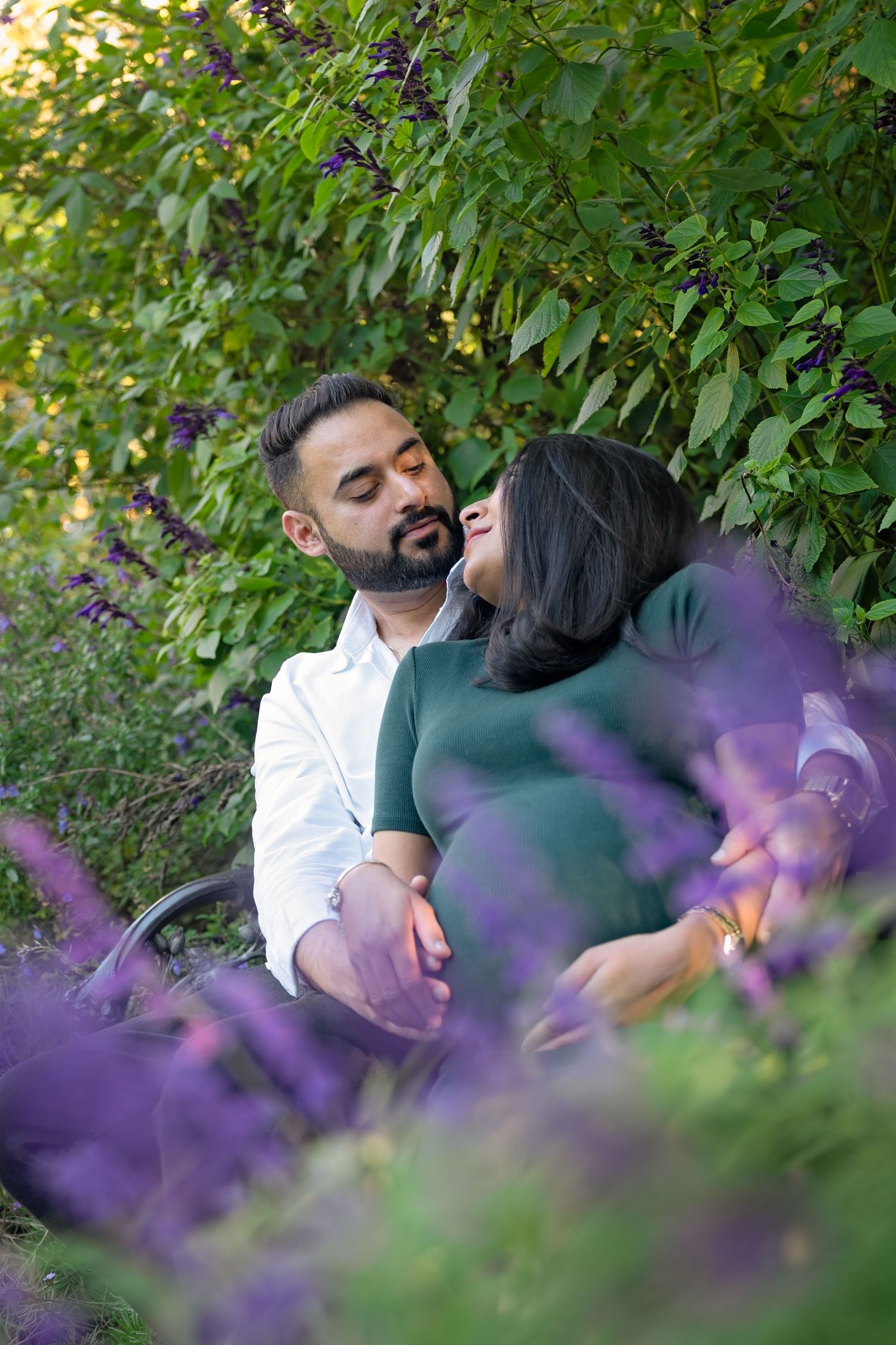 A romantic couple shares tender moments among lush green foliage and blooming purple flowers in a garden setting.