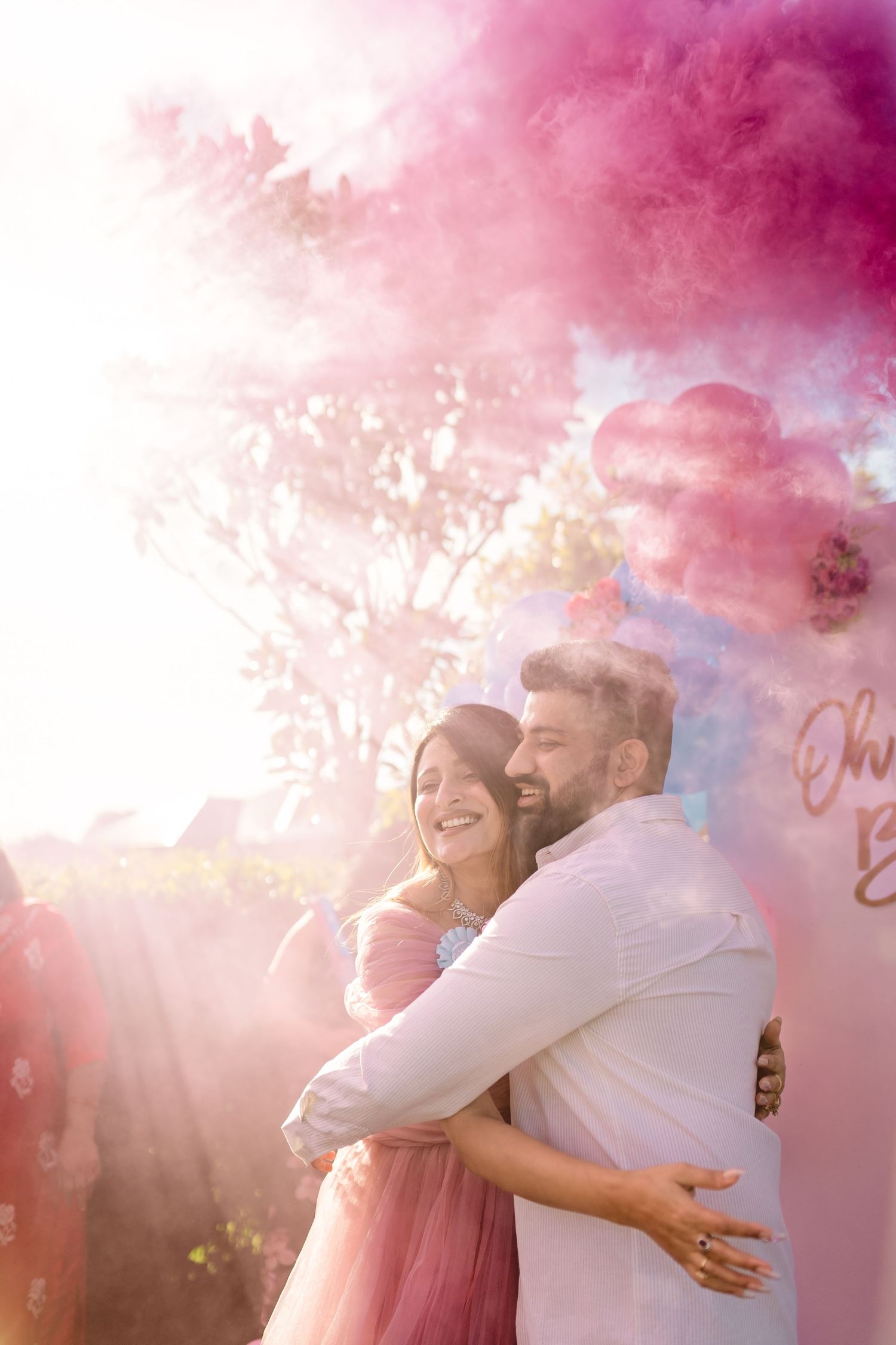 Couple embraces joyfully surrounded by pink smoke during outdoor celebration with dreamy romantic atmosphere.