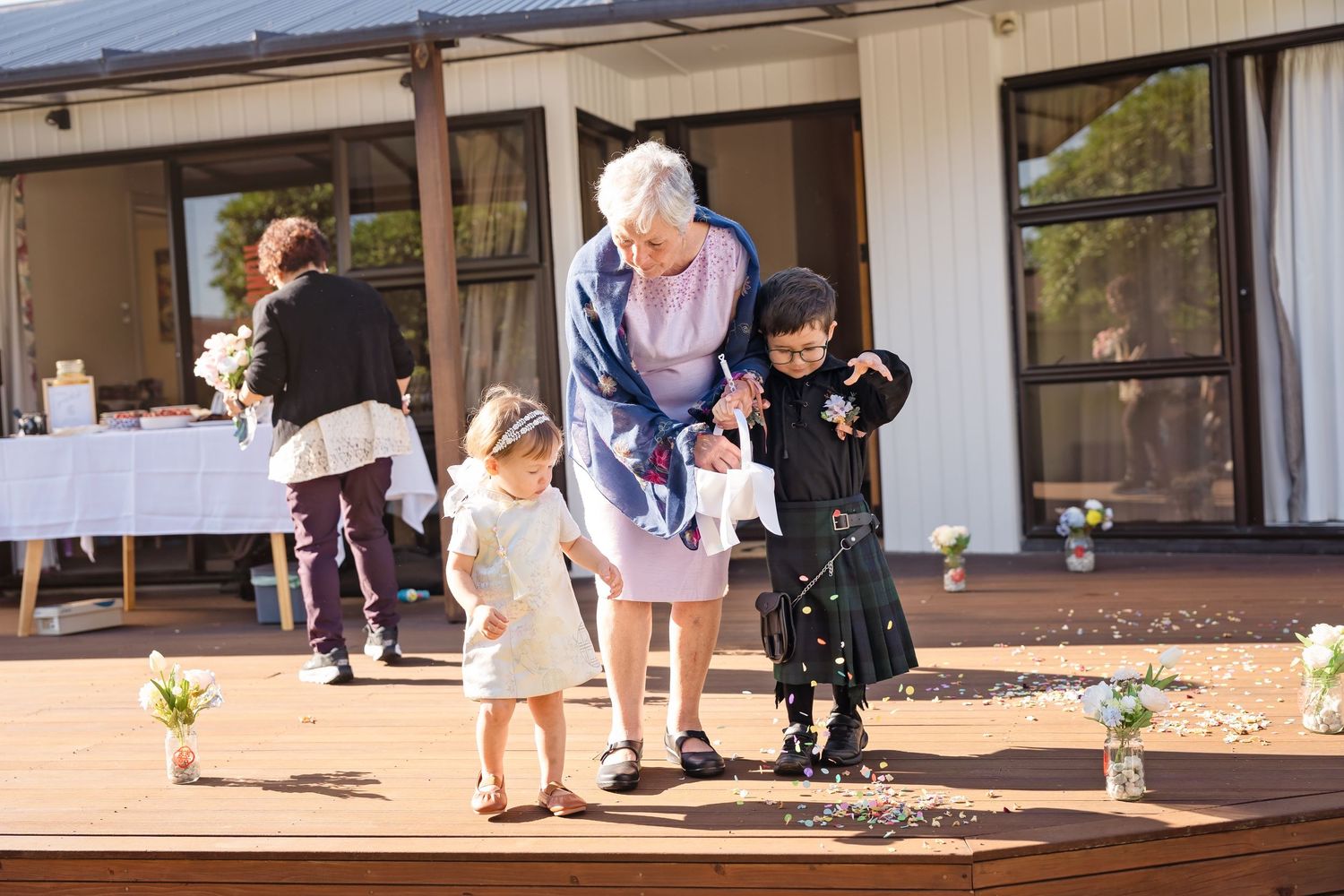 Elderly woman helps two young flower girls scatter petals on wooden deck at outdoor wedding ceremony.