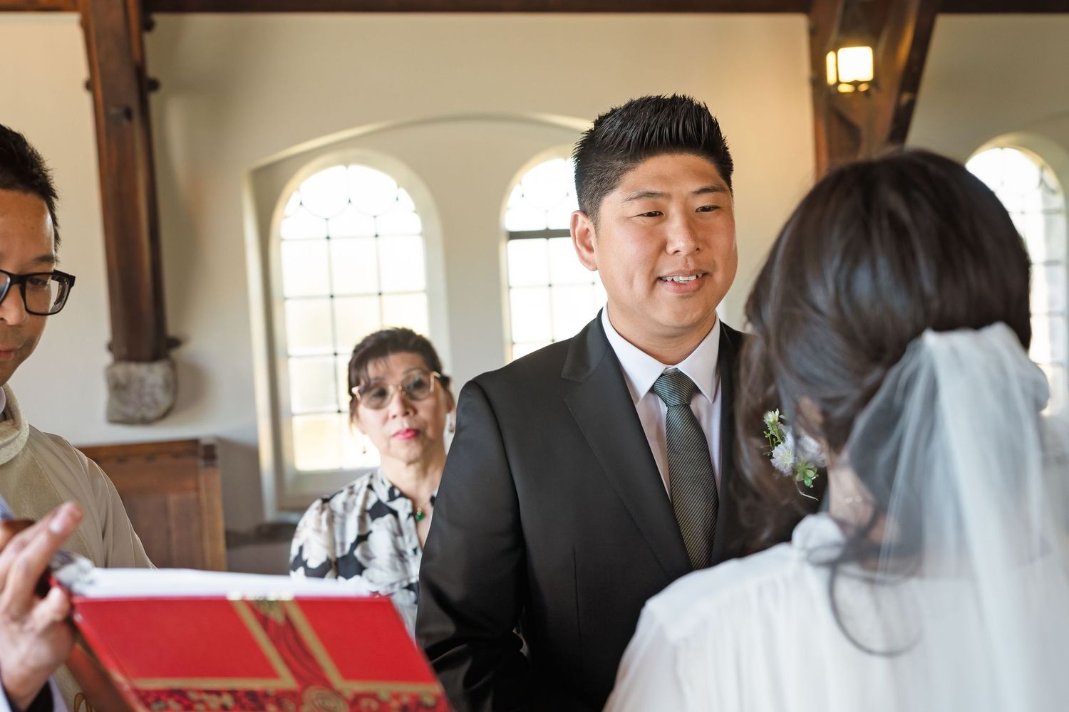 Smiling groom in dark suit faces bride during wedding ceremony inside a beautiful historic chapel.