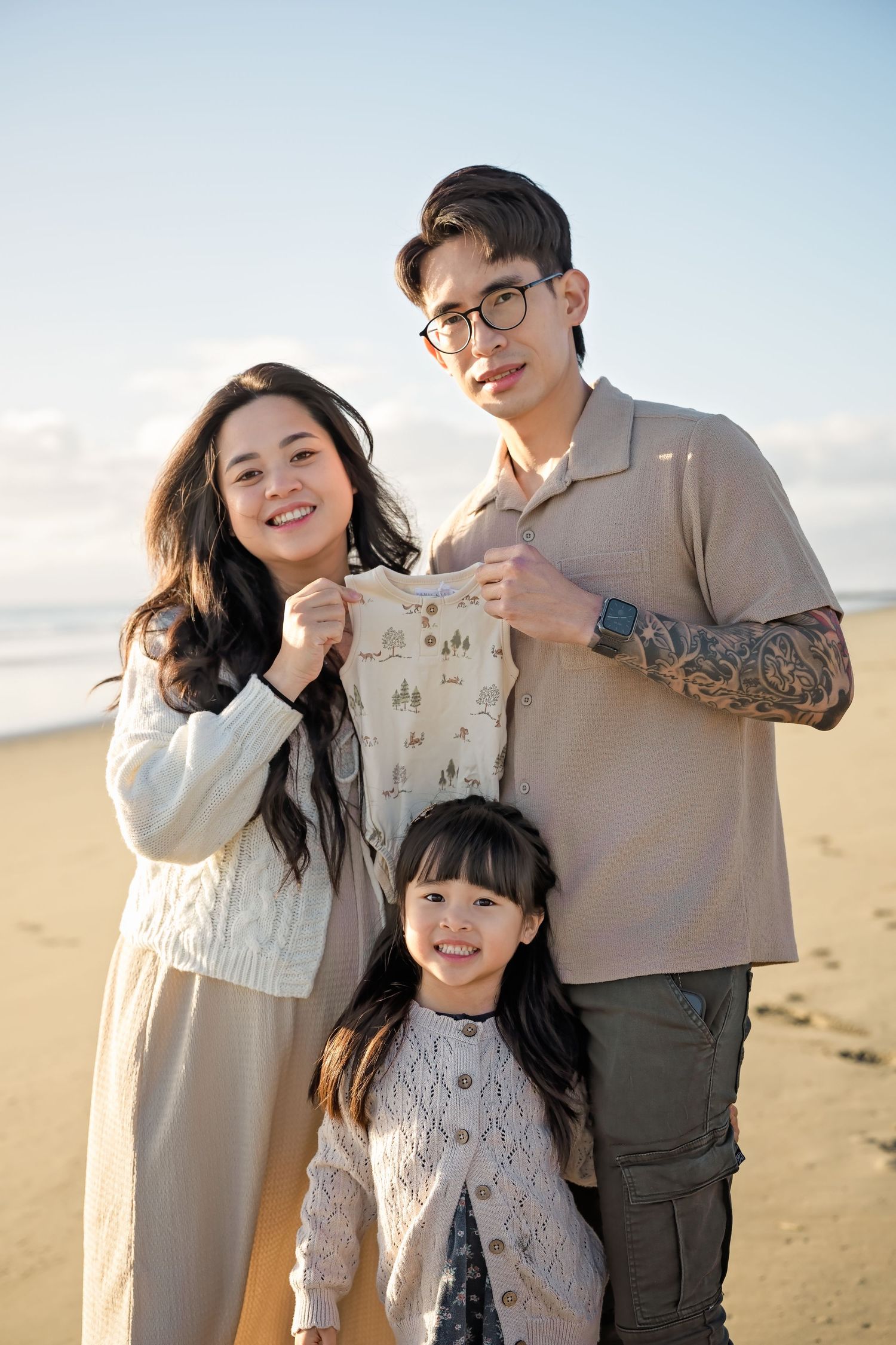 A happy Asian family of three poses on a sandy beach, parents holding a baby onesie for a pregnancy announcement.
