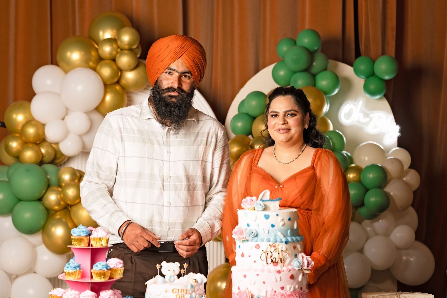 Sikh couple in matching orange outfits pose with tiered baby shower cake and balloon garland backdrop.