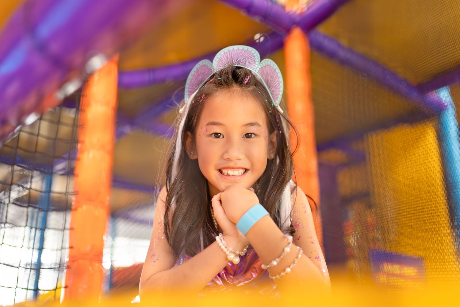 Smiling young girl with mouse ears headband playing in colorful indoor playground equipment.