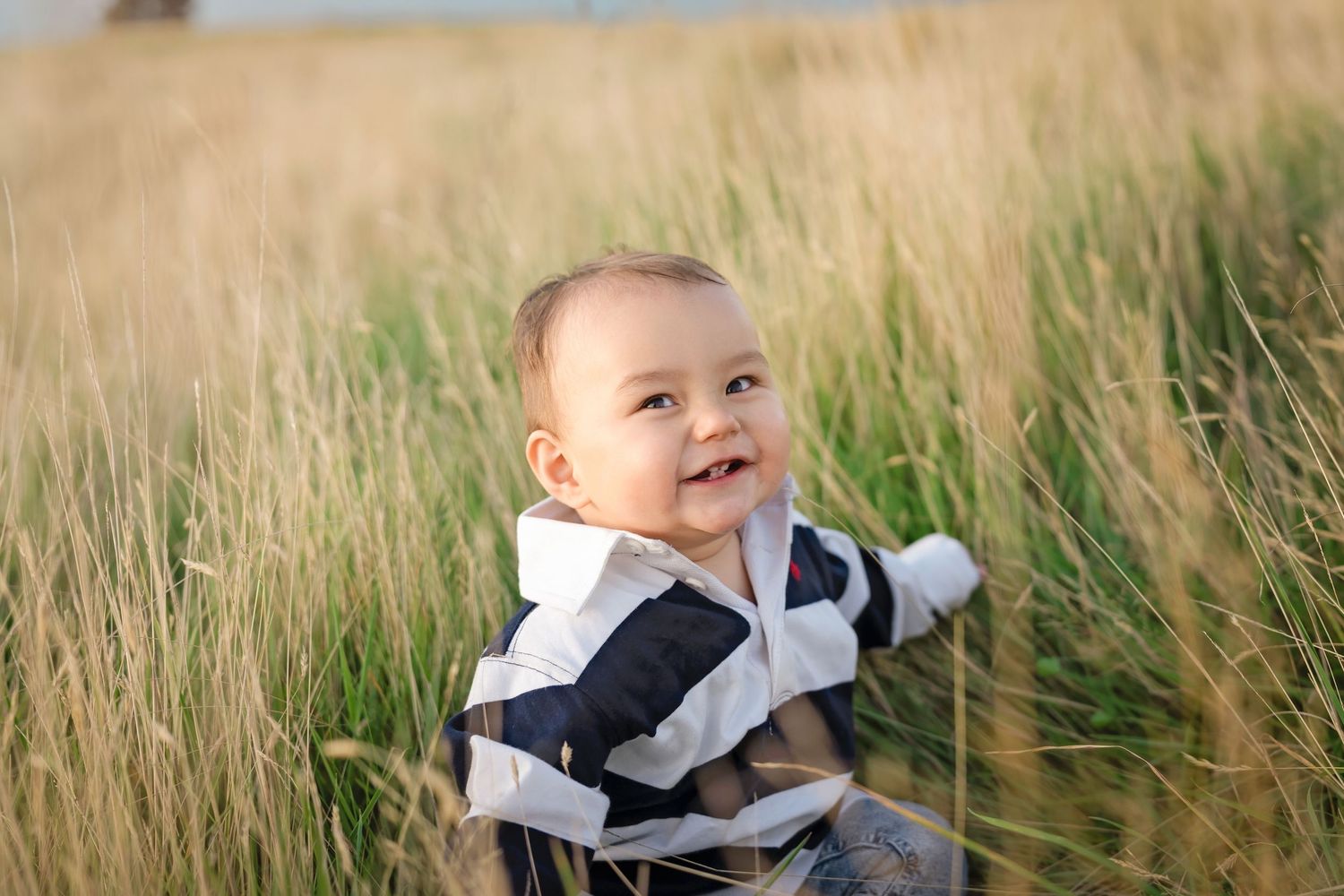 Happy baby sitting in tall golden grass field, smiling joyfully while wearing a striped outfit outdoors.