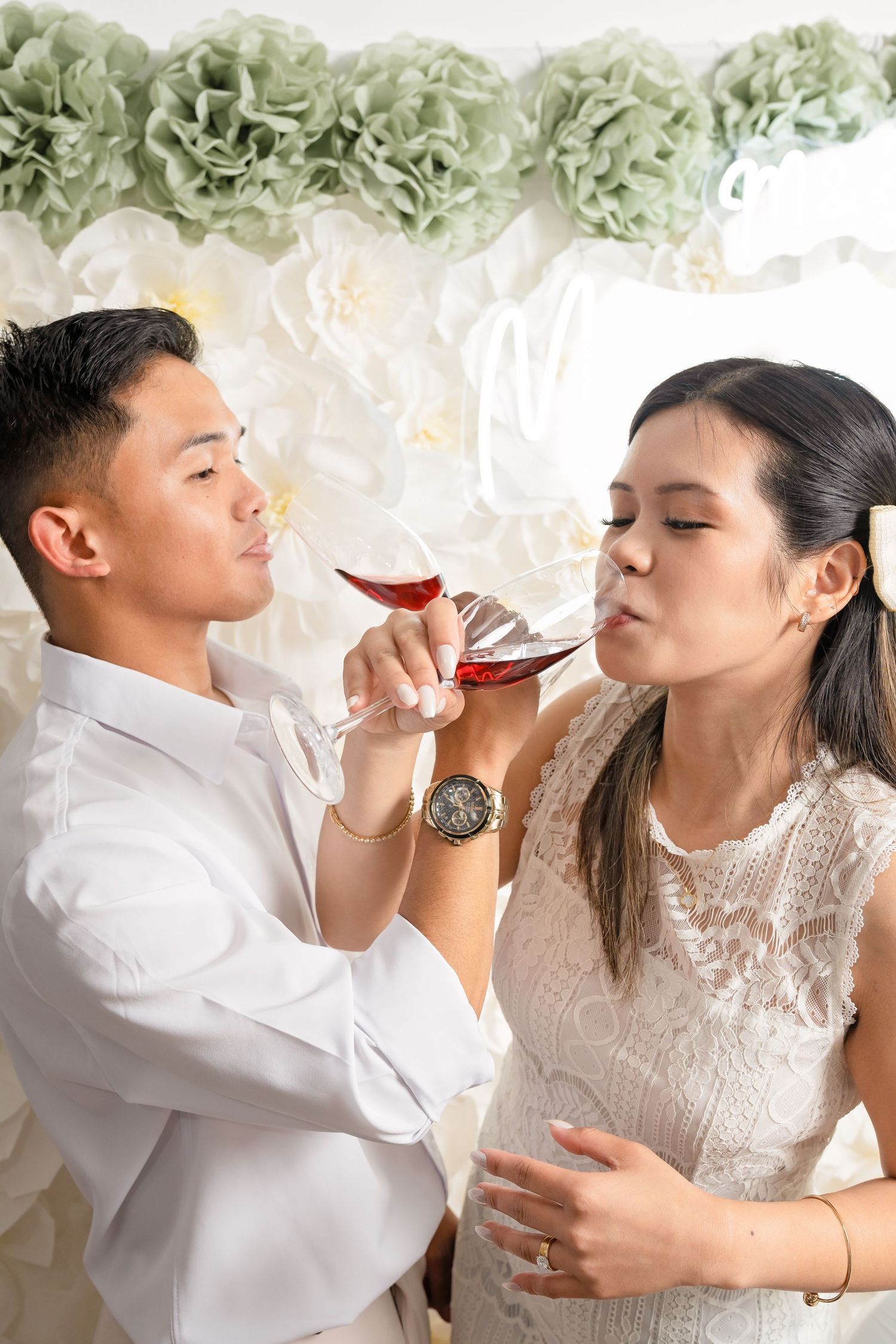 Asian couple in wedding attire sharing a glass of red wine, with floral decorations in the background.