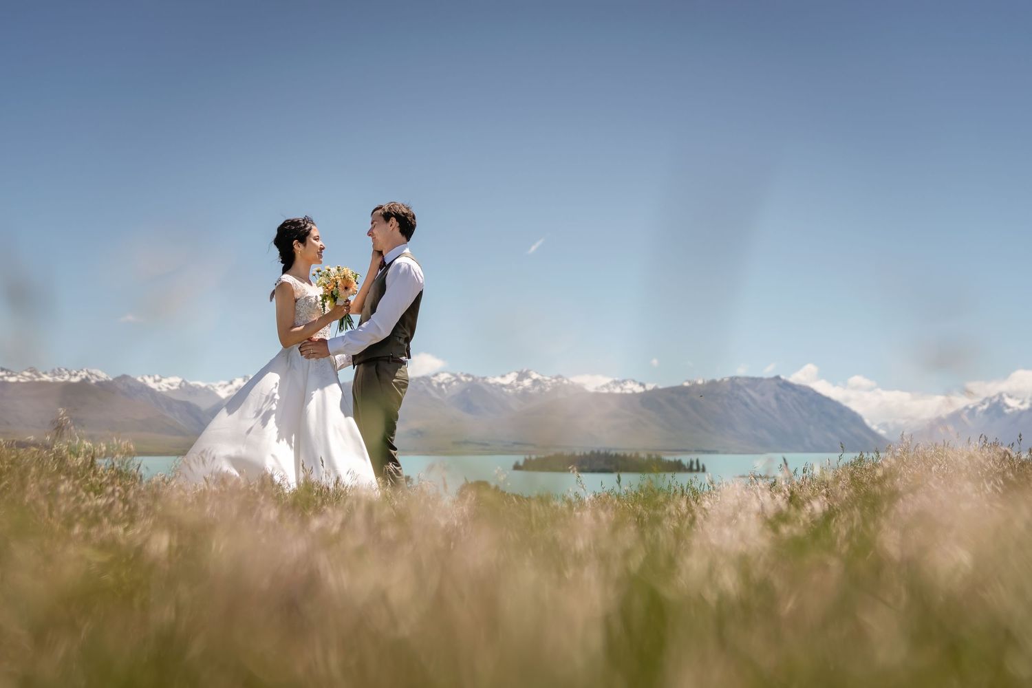 Bride and groom embracing in a golden field with a stunning lake and snow-capped mountains behind.