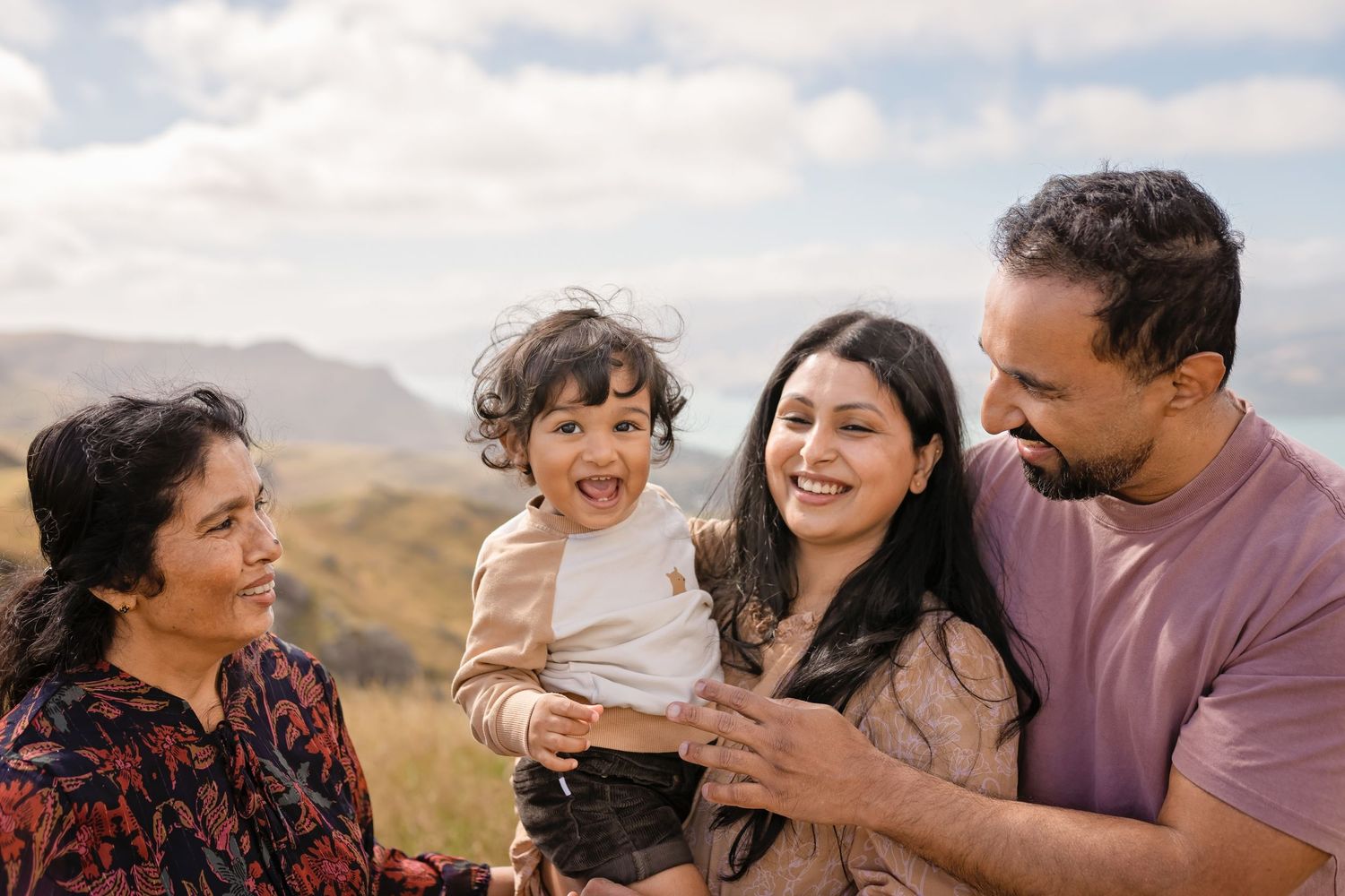 Happy South Asian family with grandmother, parents, and toddler laughing outdoors on a scenic hillside.
