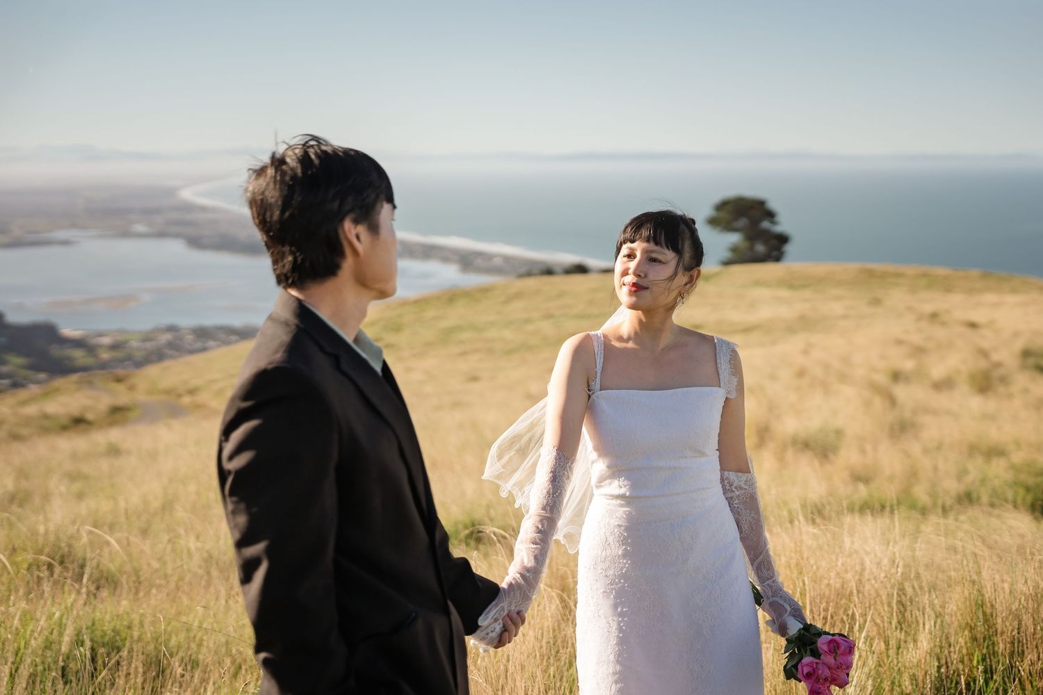 Couple in wedding attire holding hands on a scenic hilltop with ocean views on a sunny day.