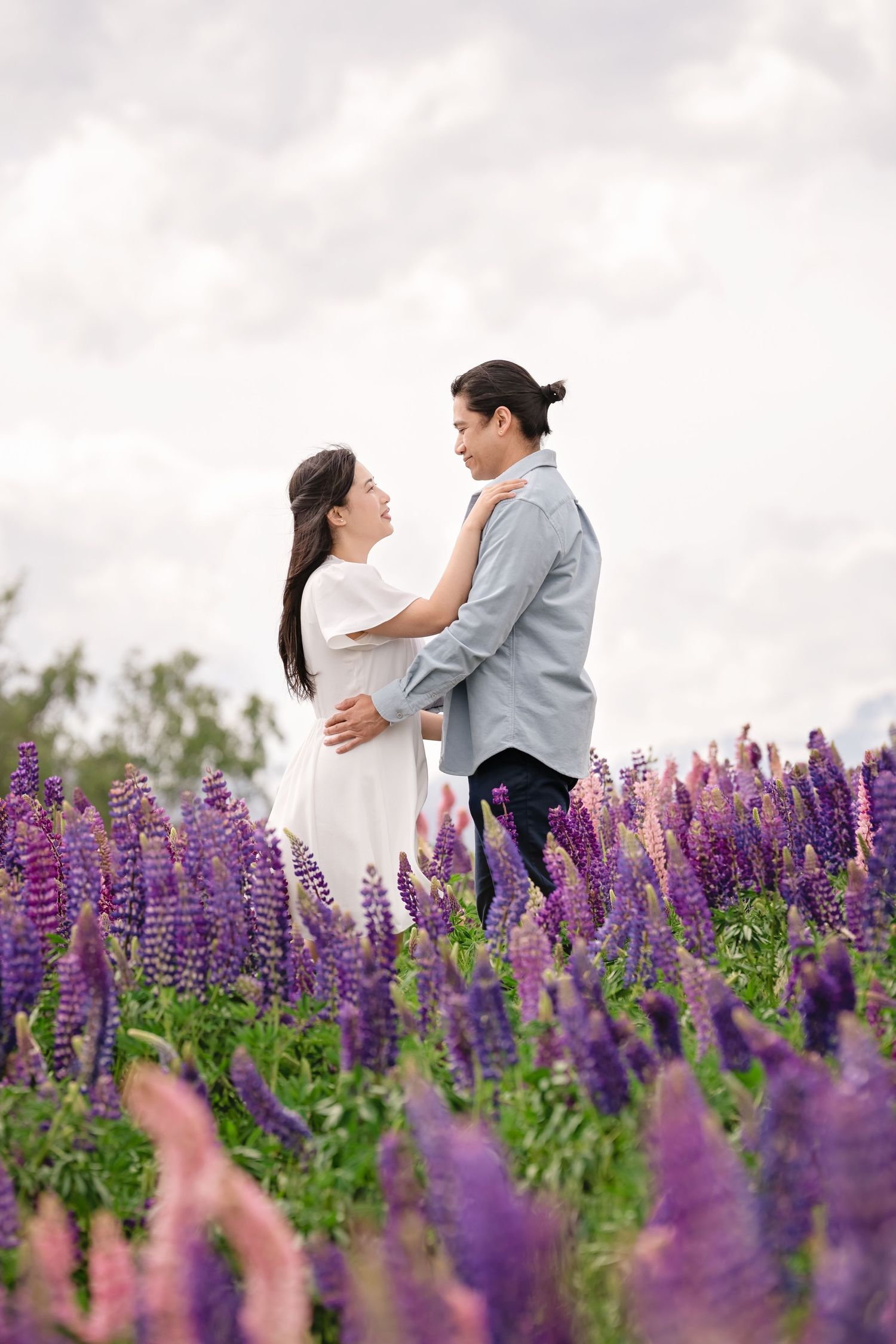 Couple embracing romantically in a vibrant purple lupine flower field under a cloudy sky.