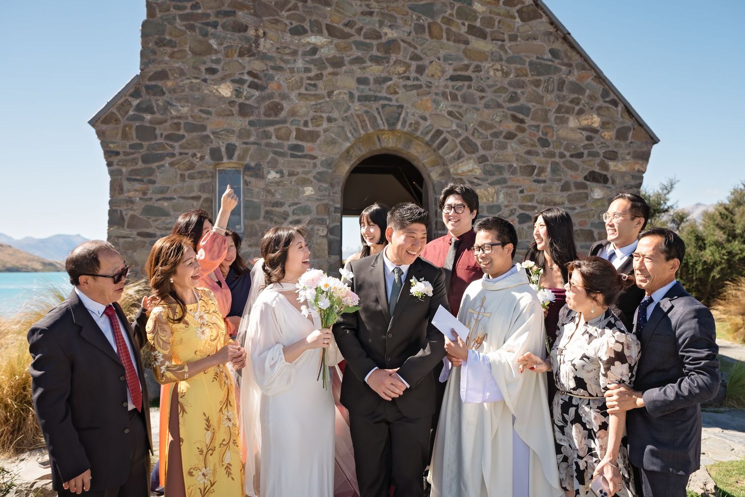 Wedding party celebrates outside historic stone chapel with stunning blue lake and mountains in New Zealand.