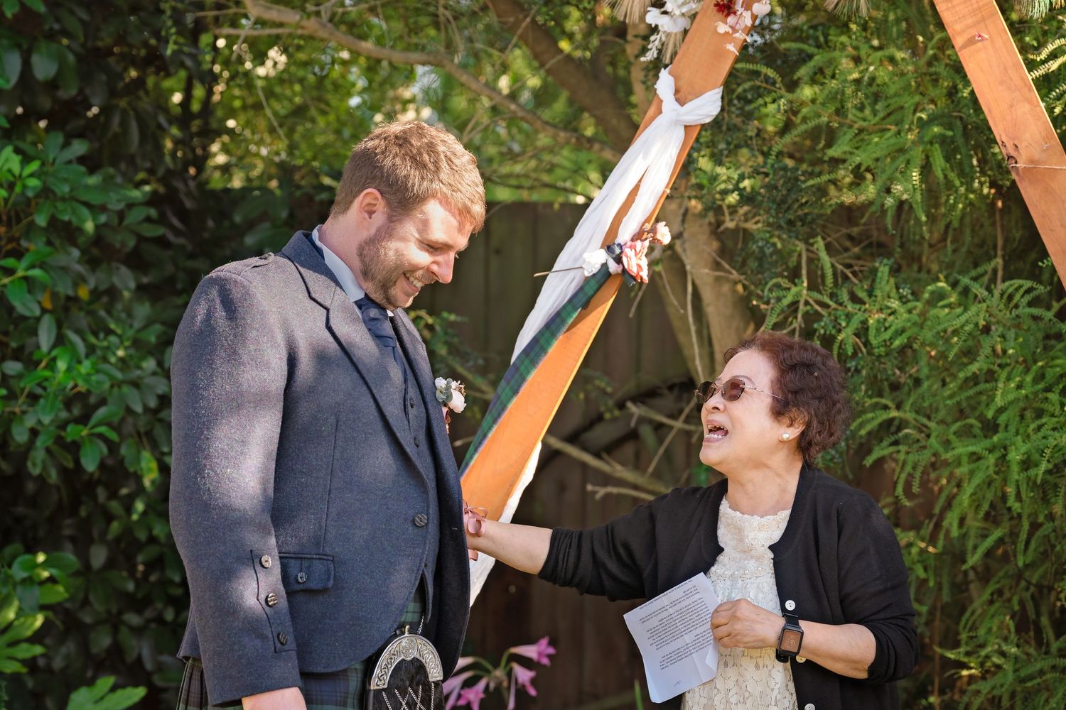 A groom in a gray suit listens as a female officiant reads from papers during an outdoor wedding ceremony.