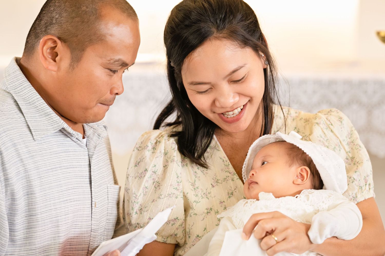 Happy Asian parents smiling at their newborn baby dressed in white, sharing a tender family moment together.