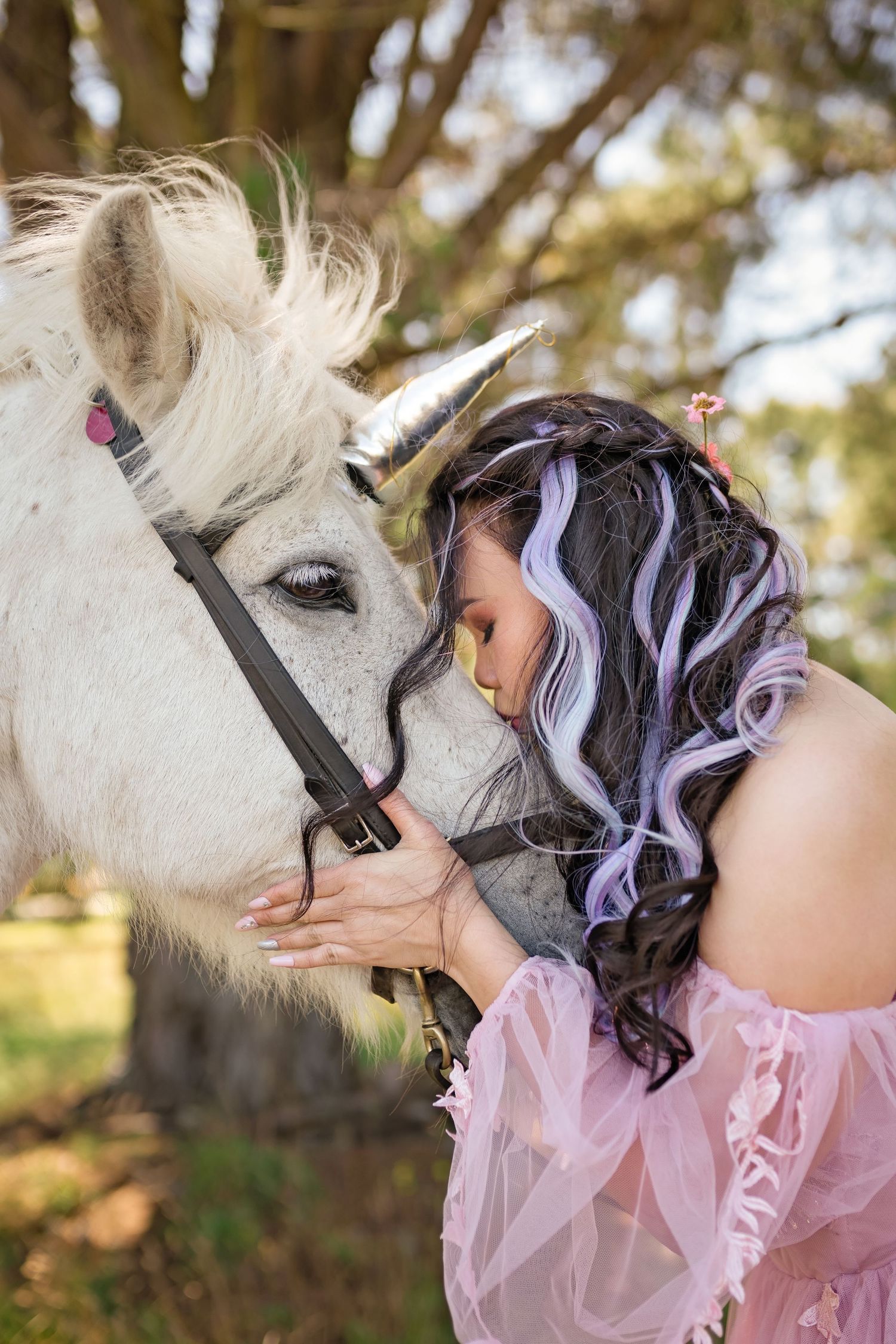 Woman with purple highlighted hair in pink dress tenderly kissing a white horse with a silver unicorn horn outdoors.
