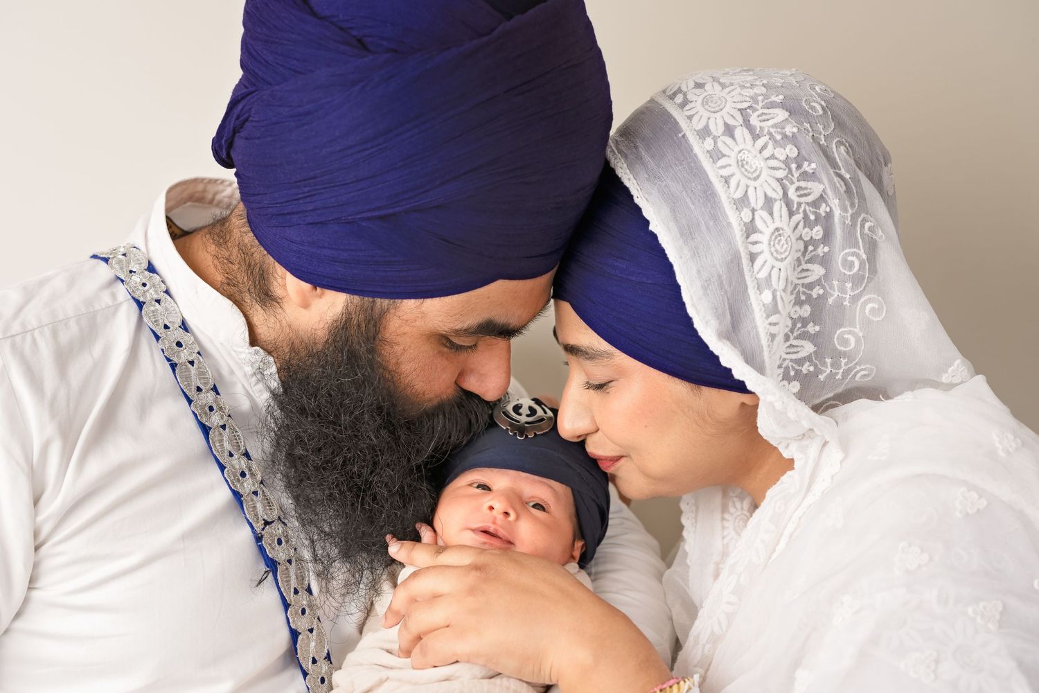 Sikh couple in traditional attire tenderly kissing their newborn baby, father wearing blue turban, mother in white dupatta.