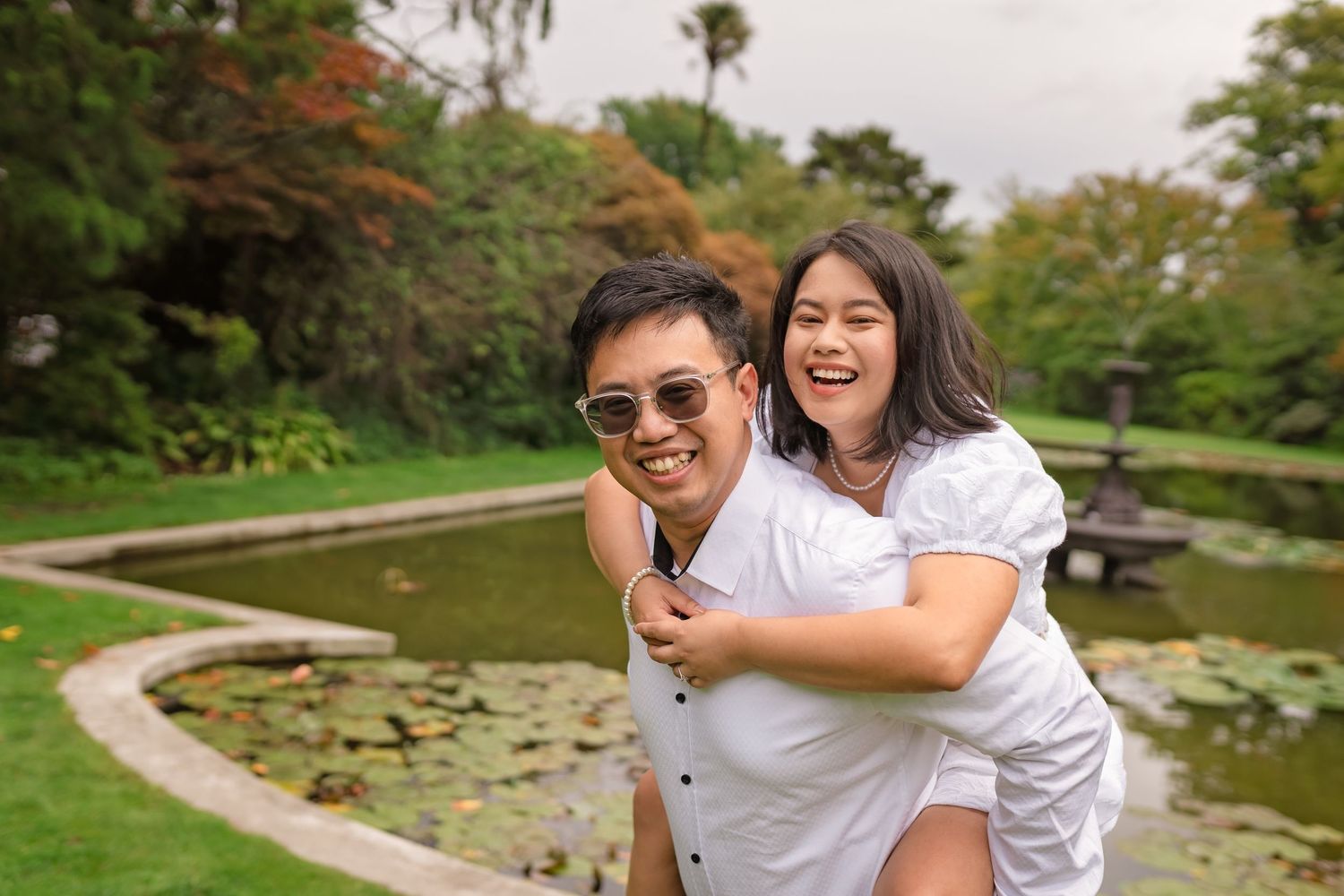Happy couple enjoying a piggyback ride in a lush garden park with a pond and lily pads in the background.