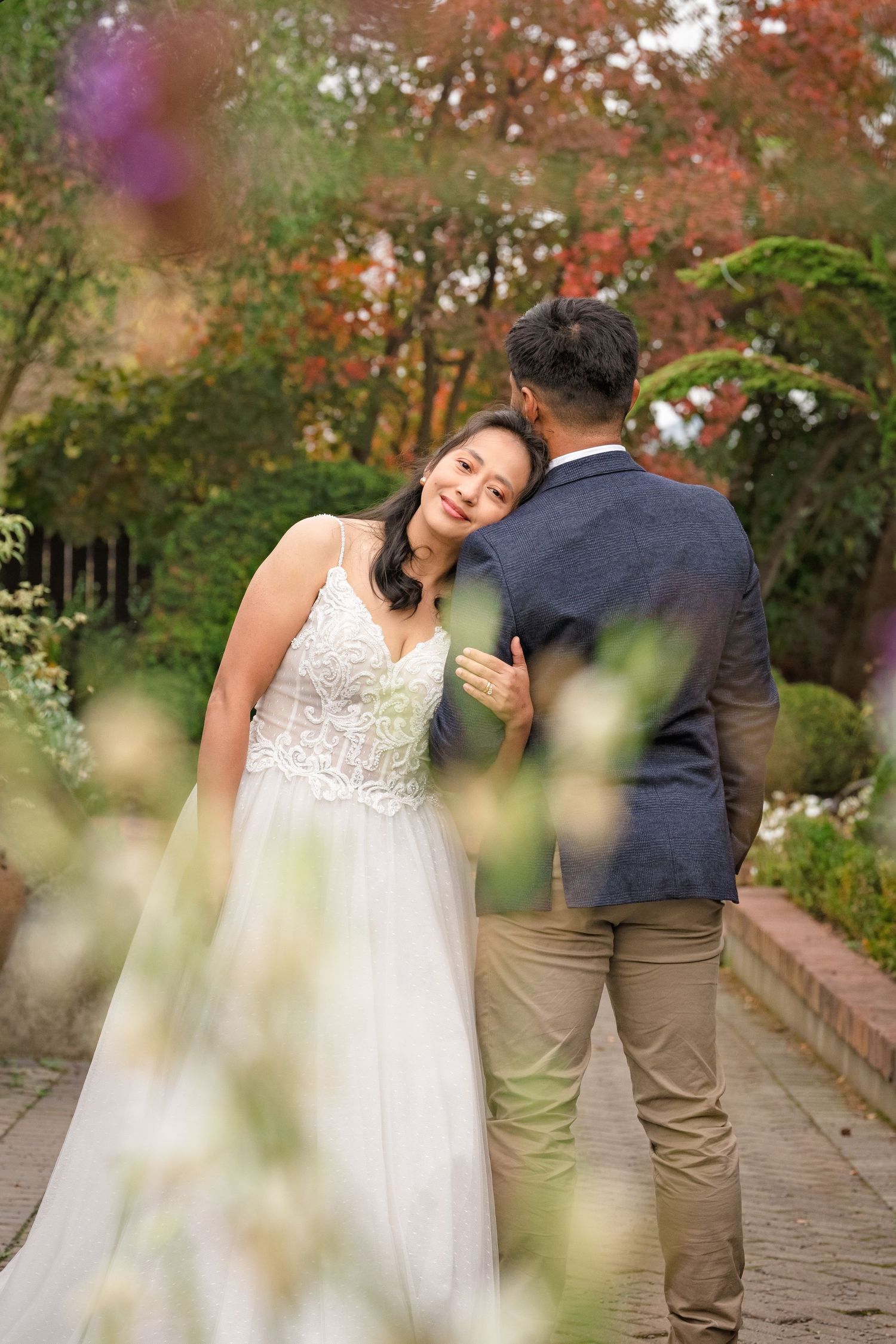 A bride in a white lace gown leans on her groom in a garden setting with colorful autumn foliage.