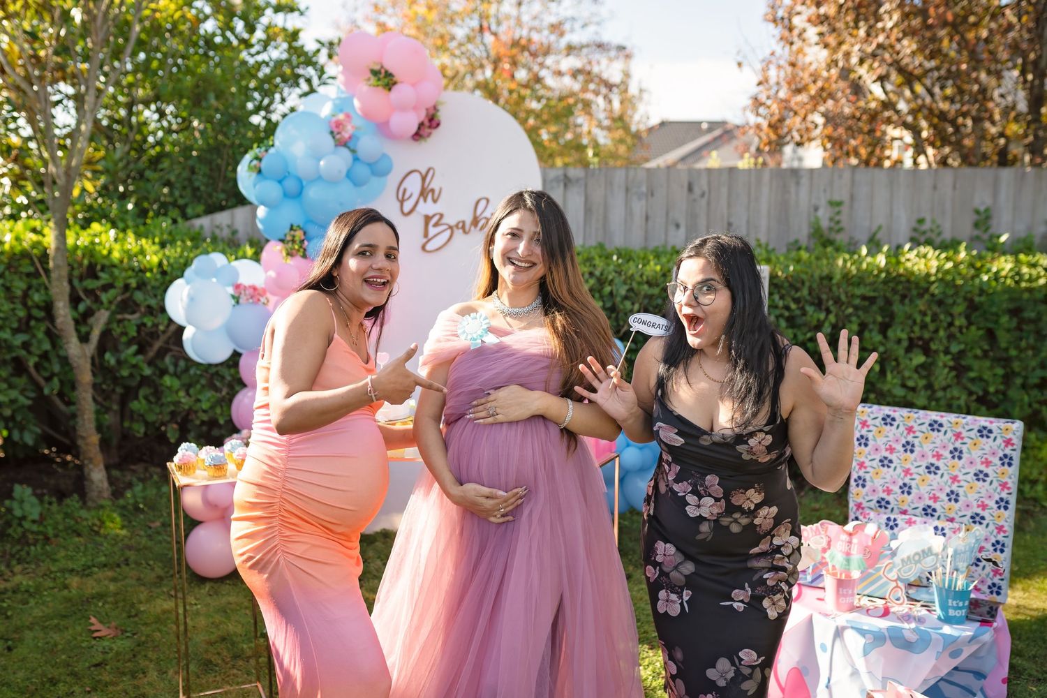 Three friends celebrate at a gender reveal party with pink and blue balloons and decorations in a backyard setting.