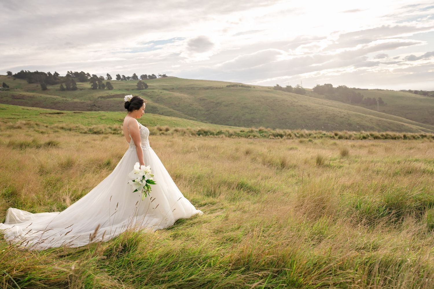 Bride in elegant white gown with long train stands in lush green rolling hills landscape holding a bouquet.