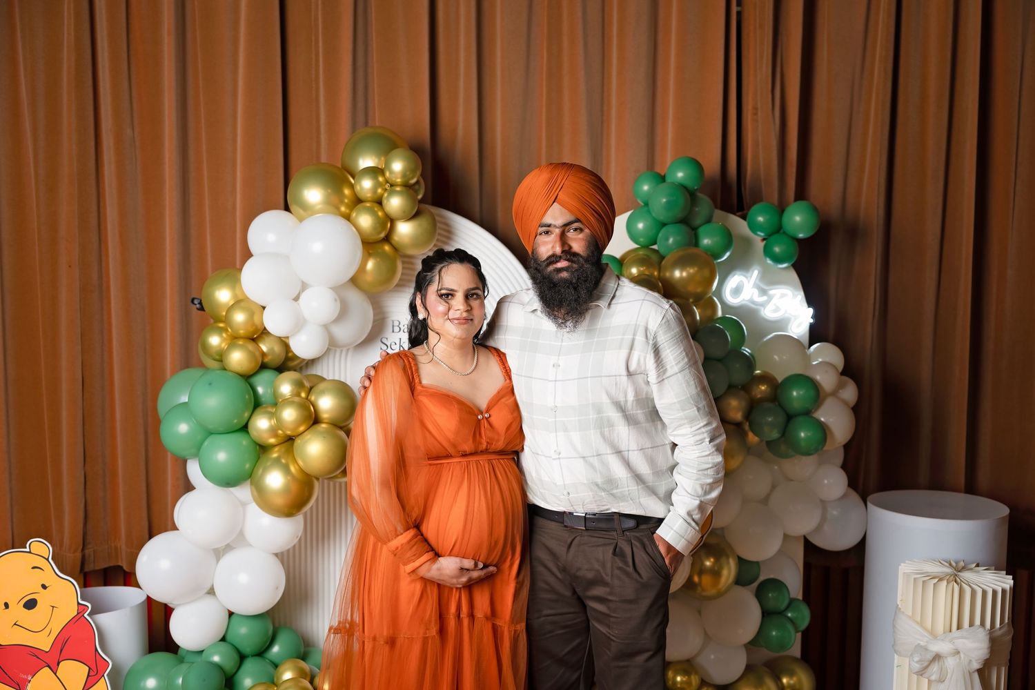 Pregnant woman in orange dress with bearded Sikh man in orange turban posing at baby shower with green and gold balloons.