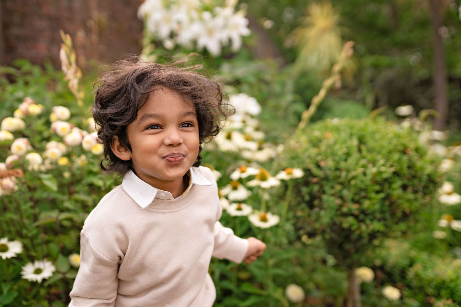 A cheerful young child with curly hair makes a funny face in a lush garden with white flowers.