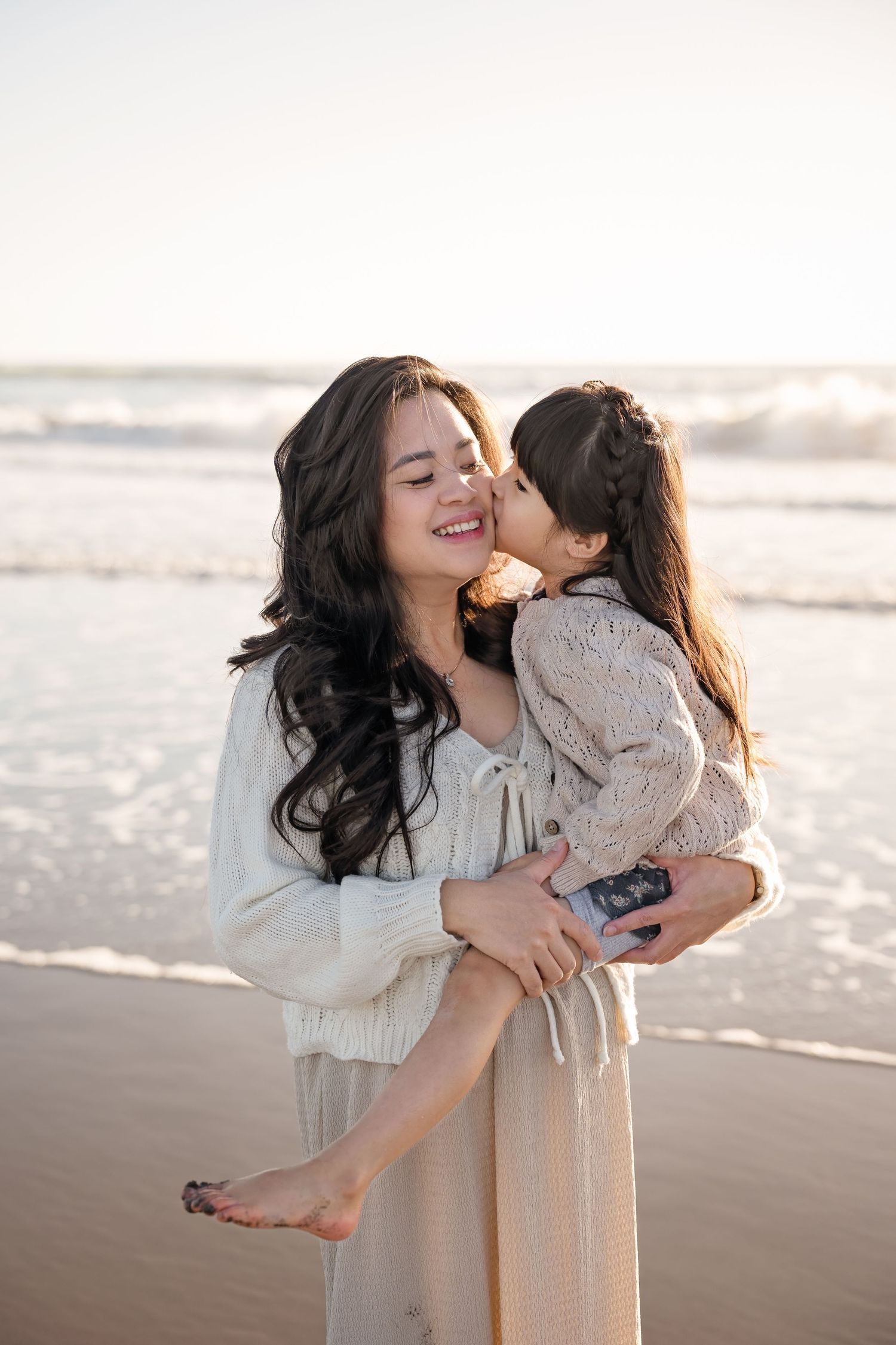 A woman holds a young girl who kisses her cheek on a sunny beach, both wearing light-colored outfits.