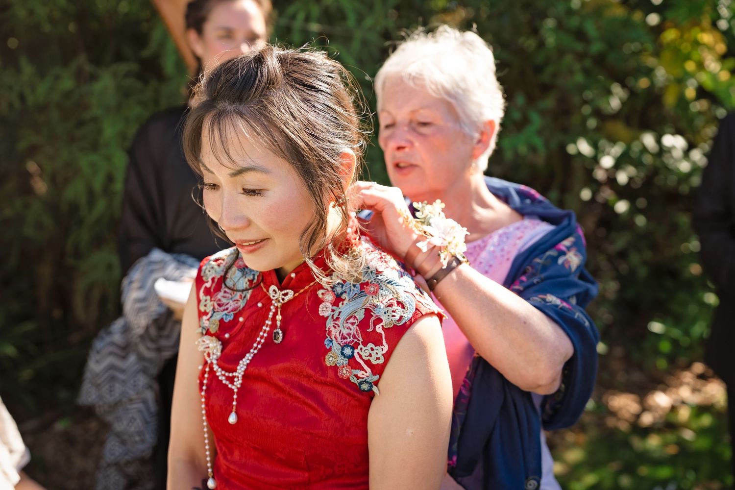 An older woman helps a bride in a red Chinese qipao dress with her jewelry at an outdoor wedding.
