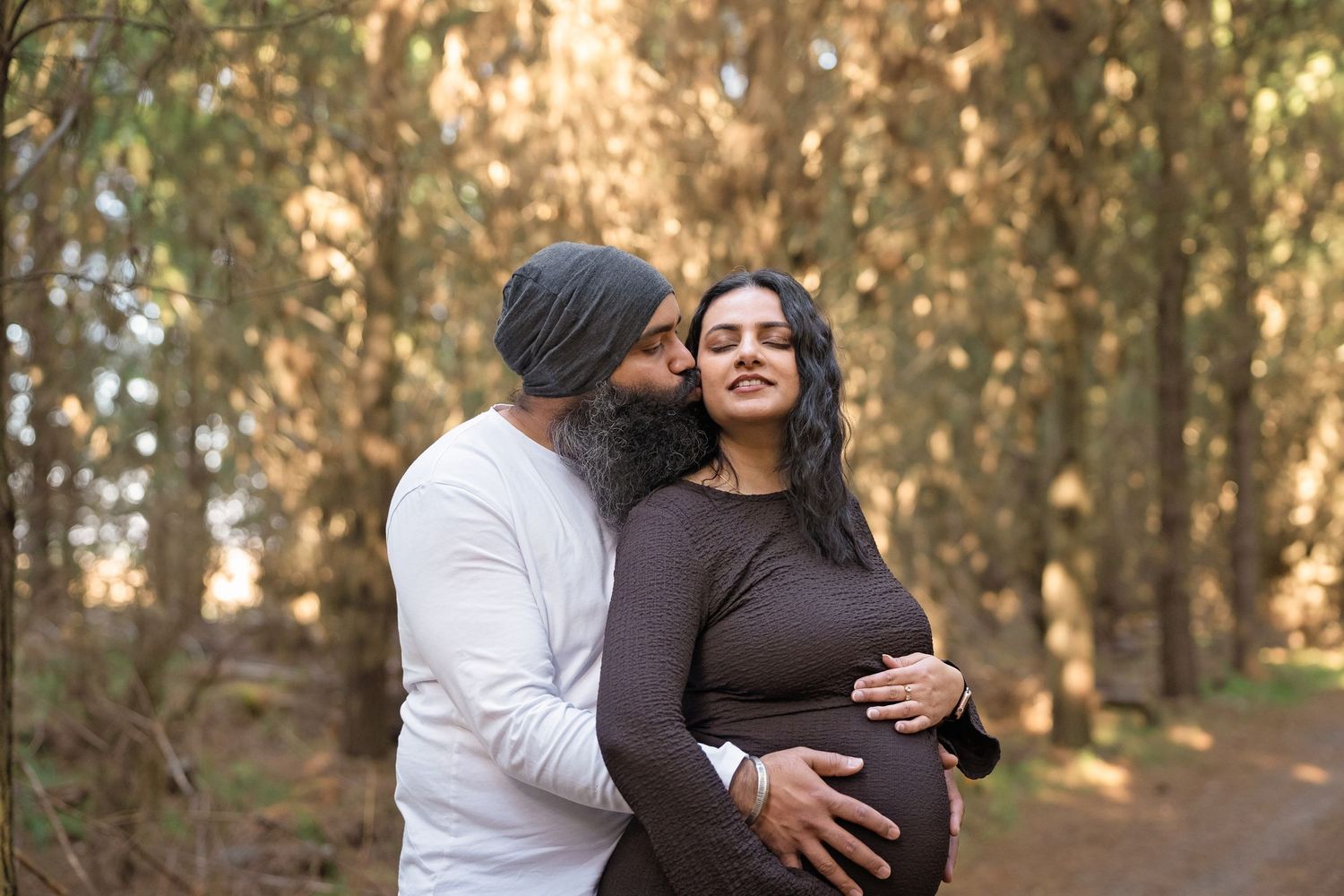 Couple sharing a tender moment in a sunlit forest, man kissing pregnant womans cheek while holding her baby bump.