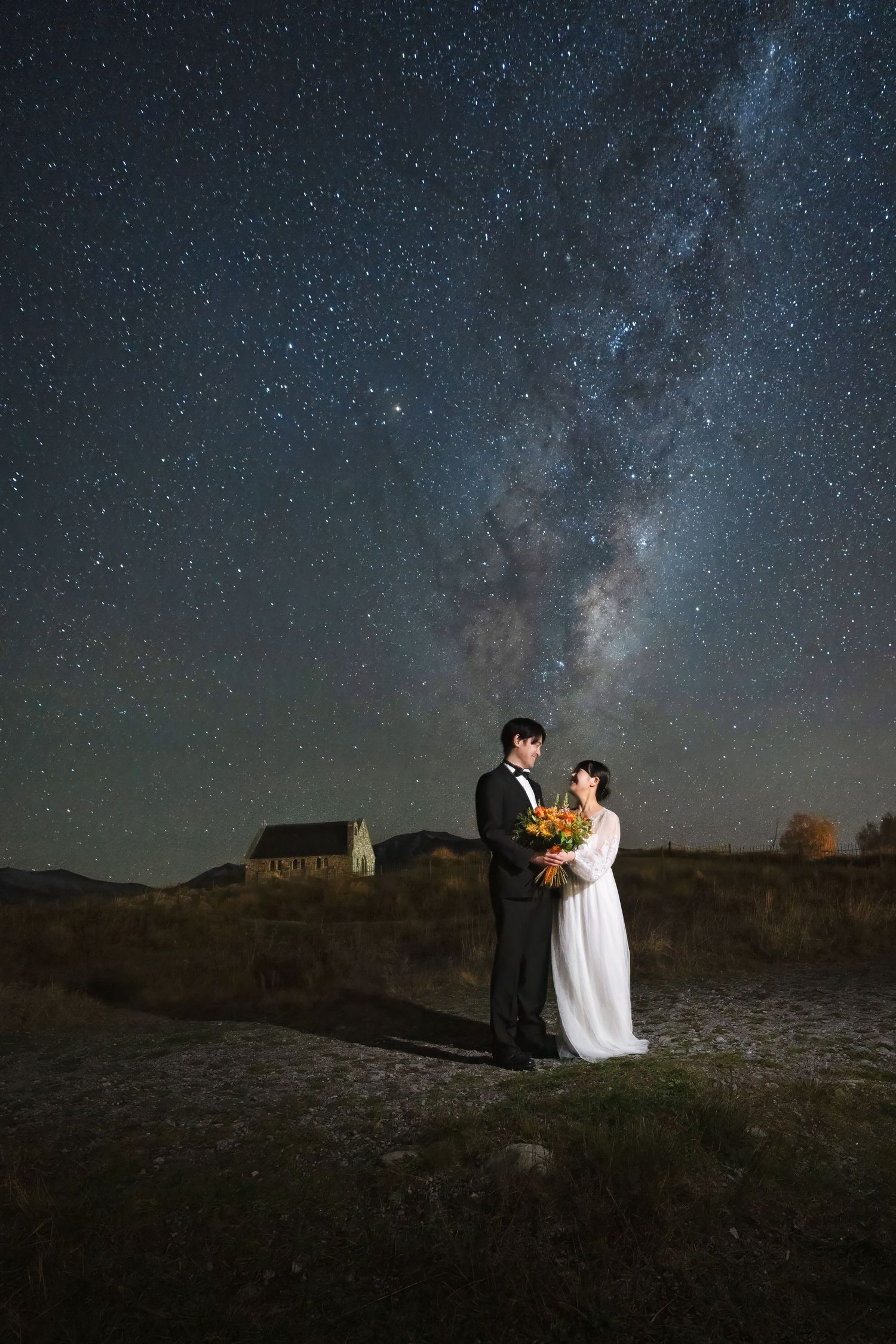 A couple shares a romantic moment under a starlit sky with the Milky Way galaxy visible above a rural landscape.