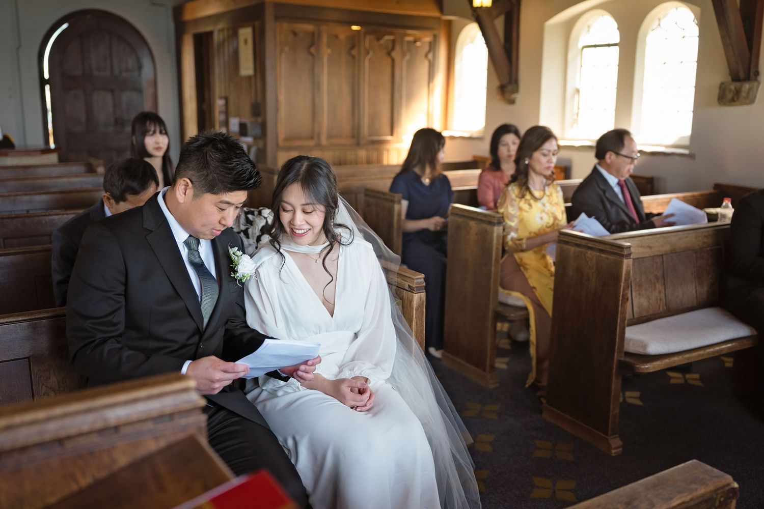Bride and groom read ceremony programs together while seated in a charming wooden church with guests.
