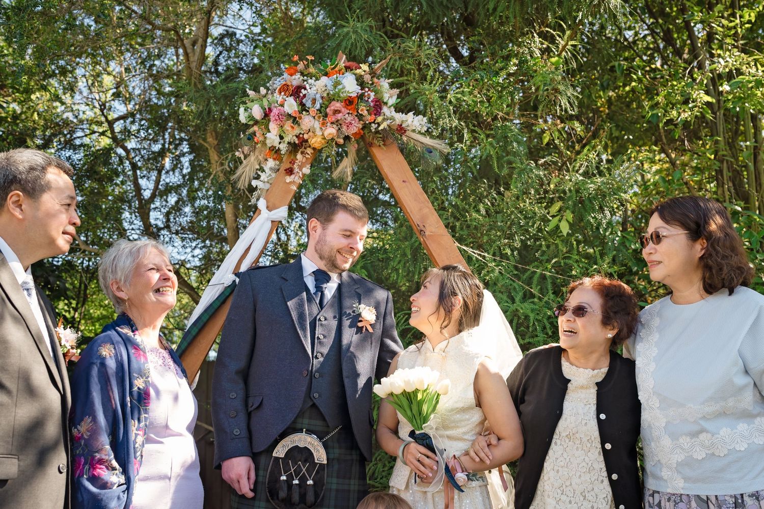 Bride and groom pose with family members under floral wedding arch in outdoor garden setting.