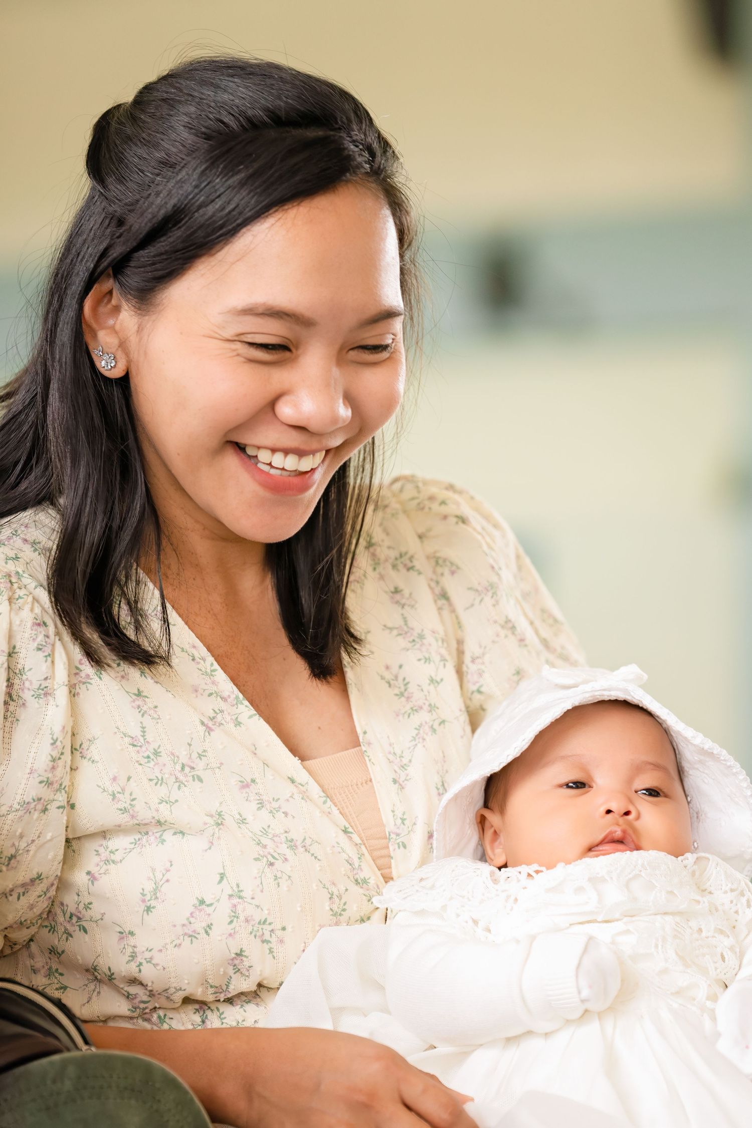 Smiling Asian mother holding newborn baby dressed in white christening outfit.