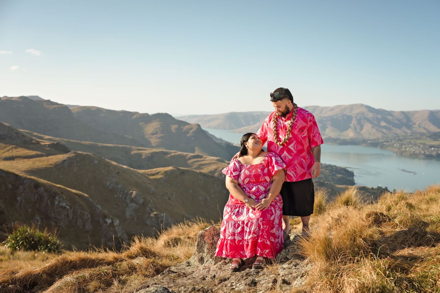 Couple in vibrant pink Pacific Island attire posing on a mountain summit with scenic harbor views in New Zealand.