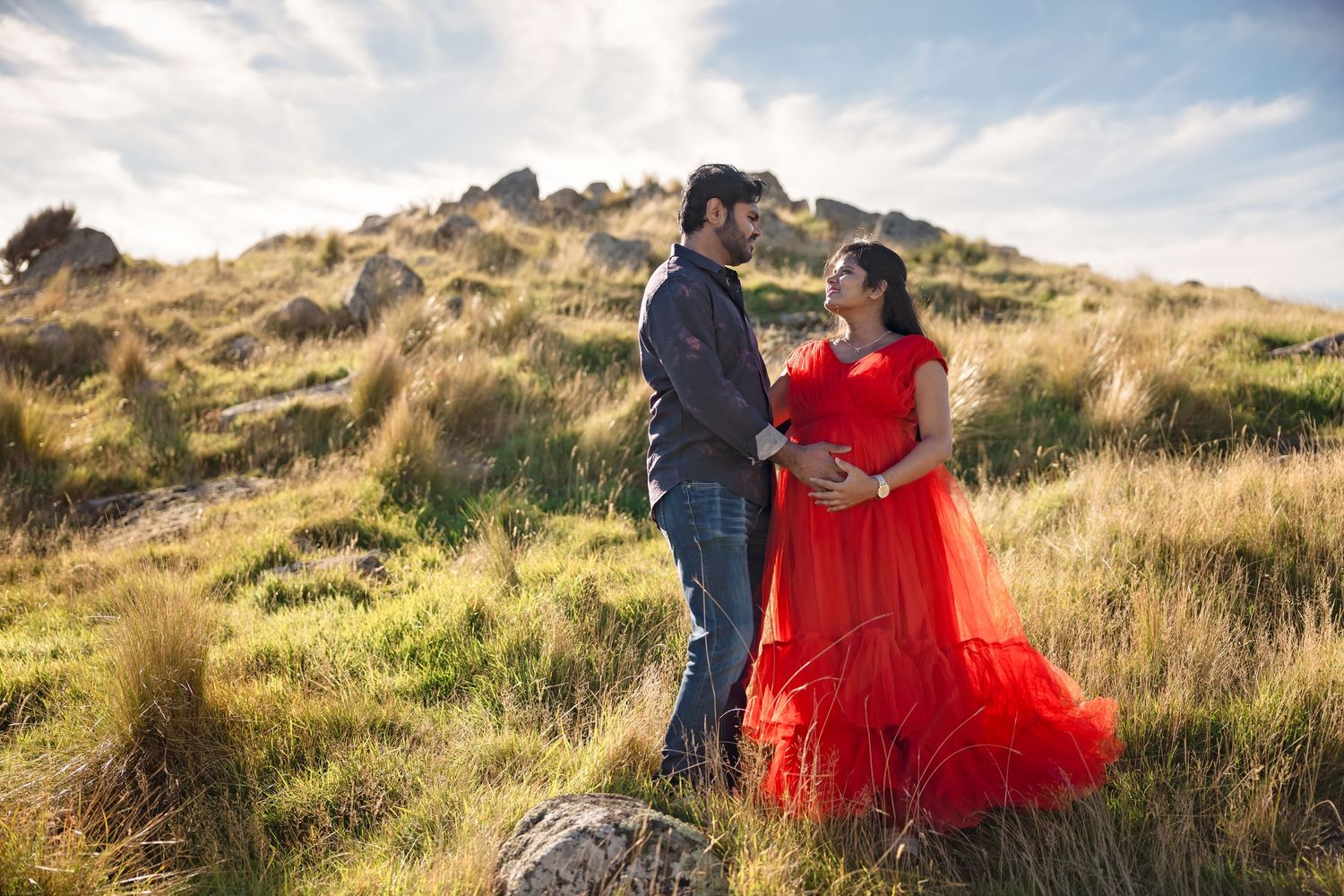 Couple in maternity photoshoot on grassy hillside, woman in flowing red gown.