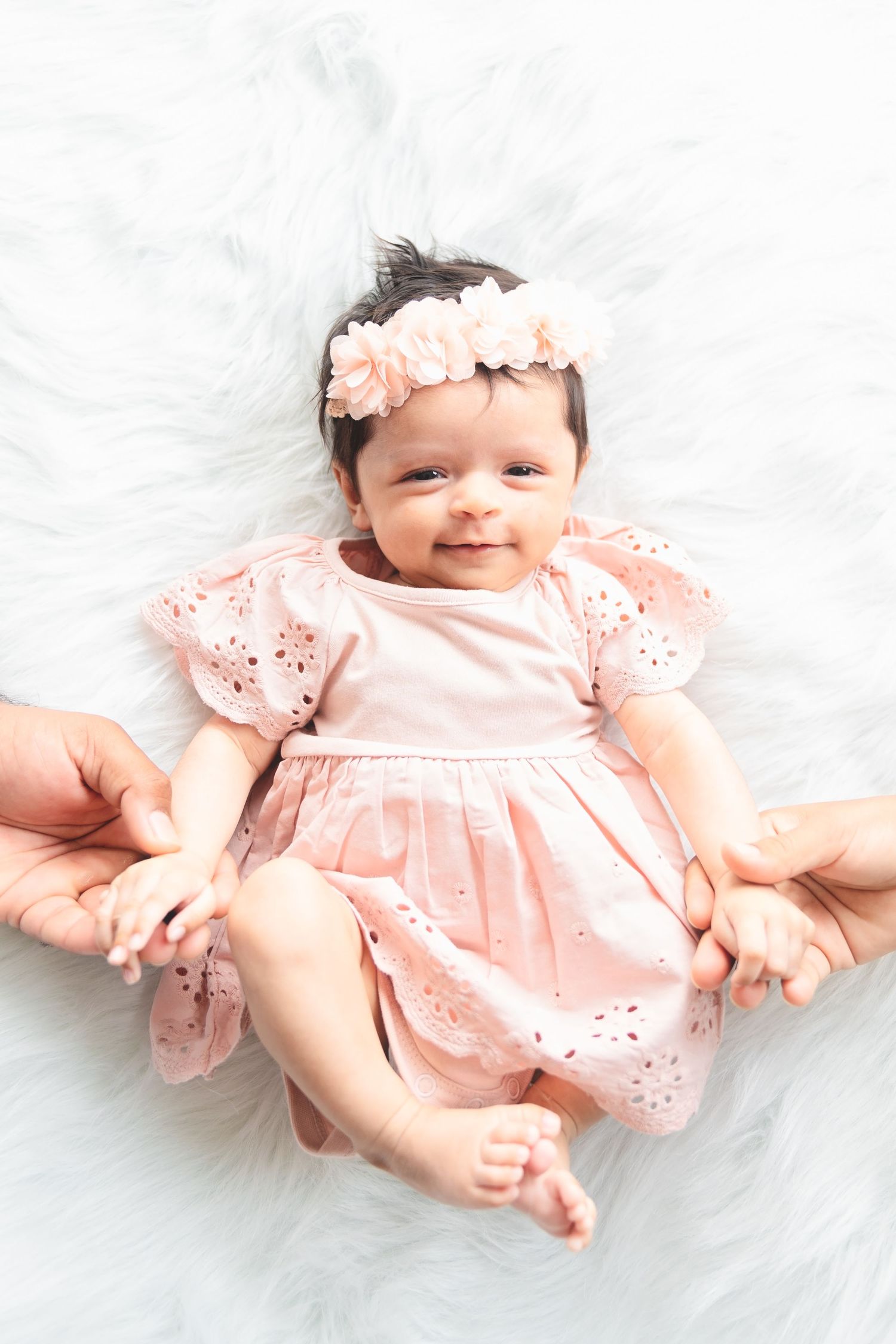Baby wearing pink eyelet dress and floral headband lying on white fluffy blanket.