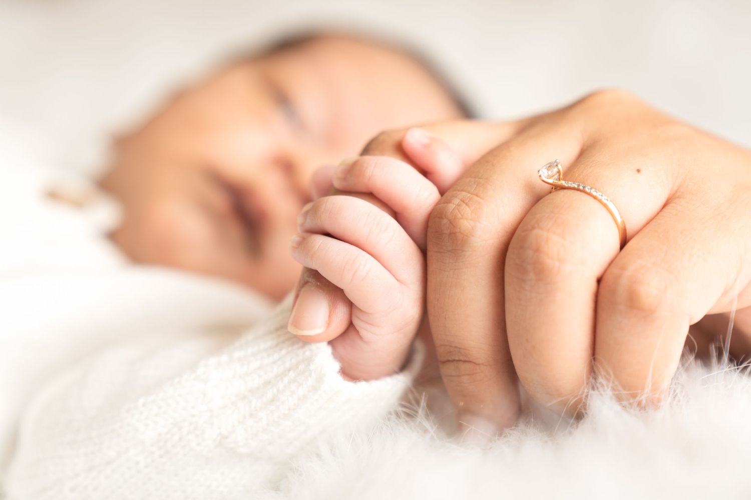 A tiny baby's hand grasps a finger with an engagement ring against a white background.