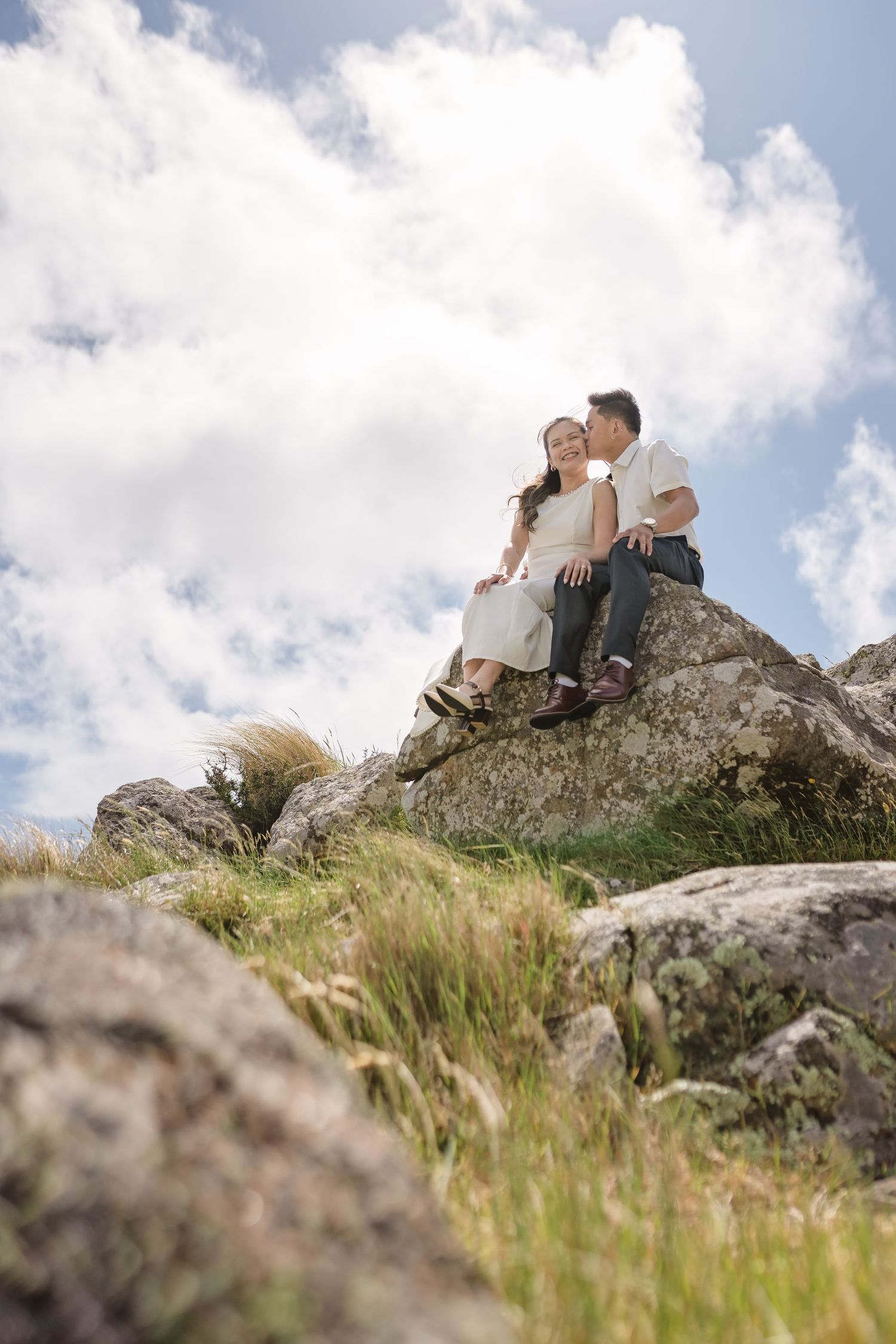 Couple sitting on rocky hilltop, man kissing woman's cheek, surrounded by green grass and cloudy blue sky.