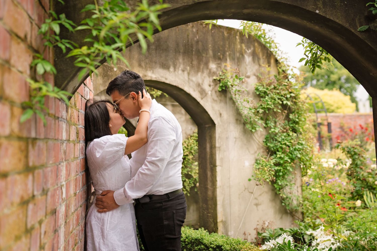 Couple sharing a romantic kiss against a brick wall beneath a stone arch surrounded by lush green ivy in a garden.