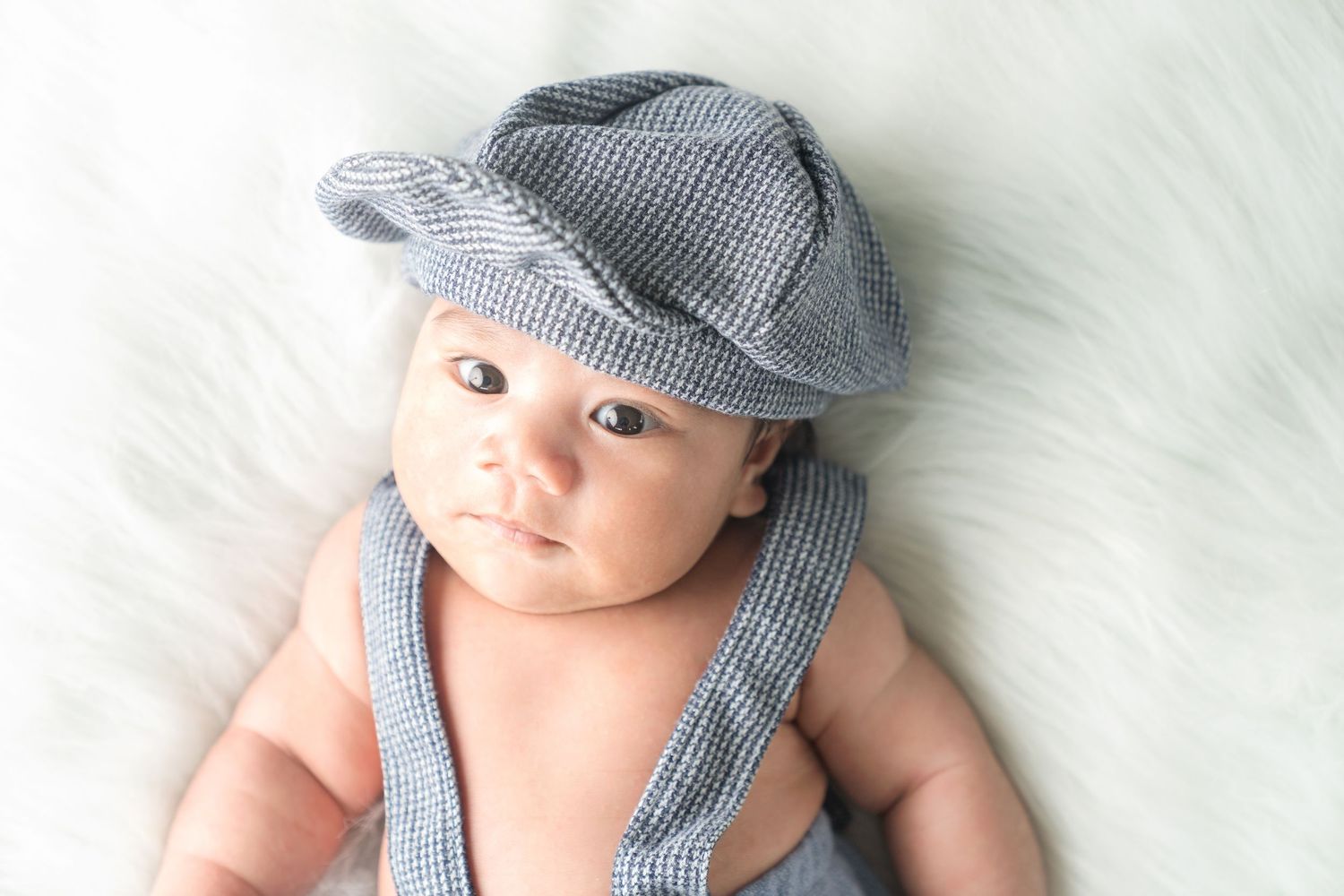 Adorable baby wearing a gray newsboy cap and striped overalls laying on soft white backdrop.