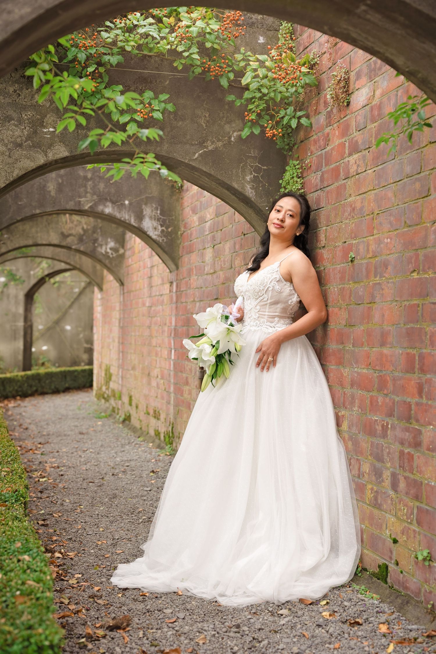 Bride in white ballgown holding calla lily bouquet poses against brick wall under stone arches in garden setting.