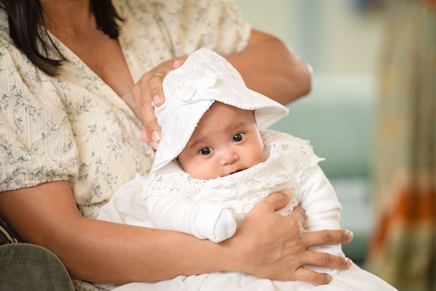 Newborn baby in white christening gown and bonnet being held by mother in floral dress.