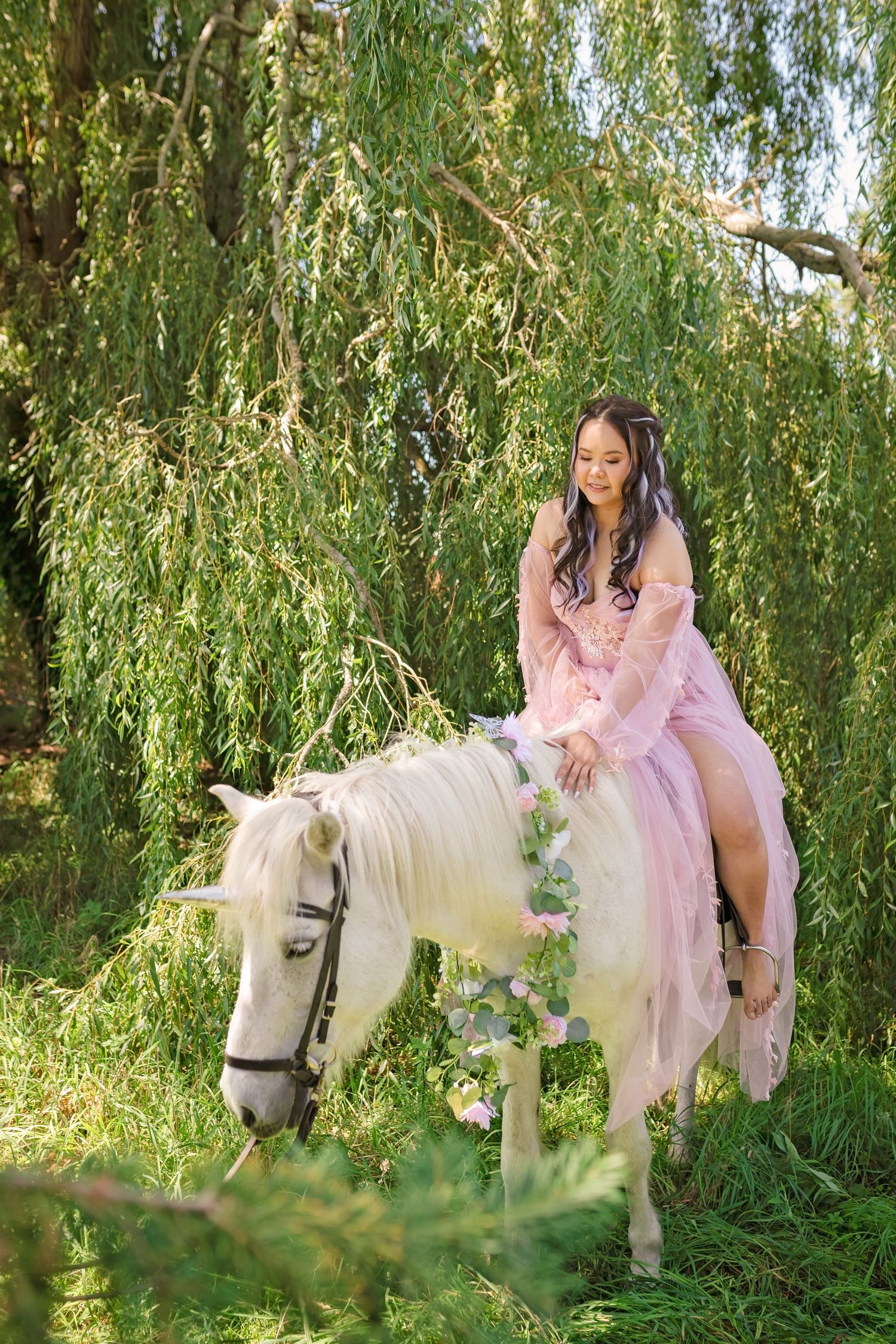 A woman in a pink gown rides a white horse adorned with flowers beneath weeping willow trees.