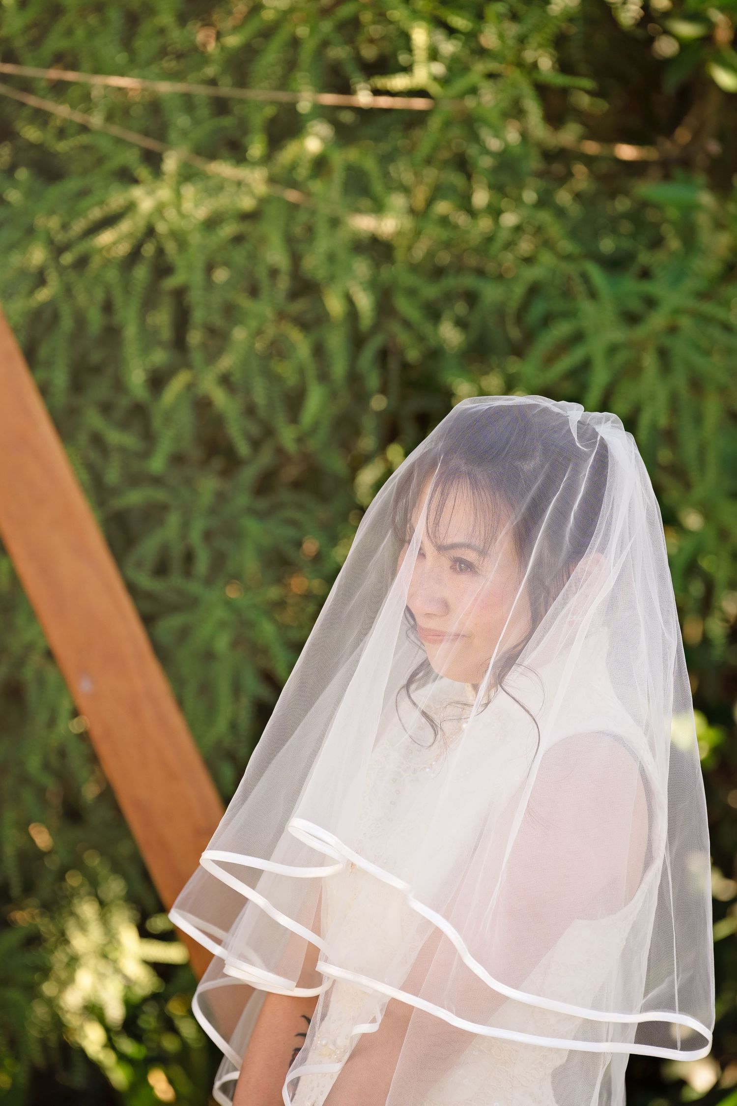 Bride wearing a white veil outdoors with lush green foliage in the background on her wedding day.