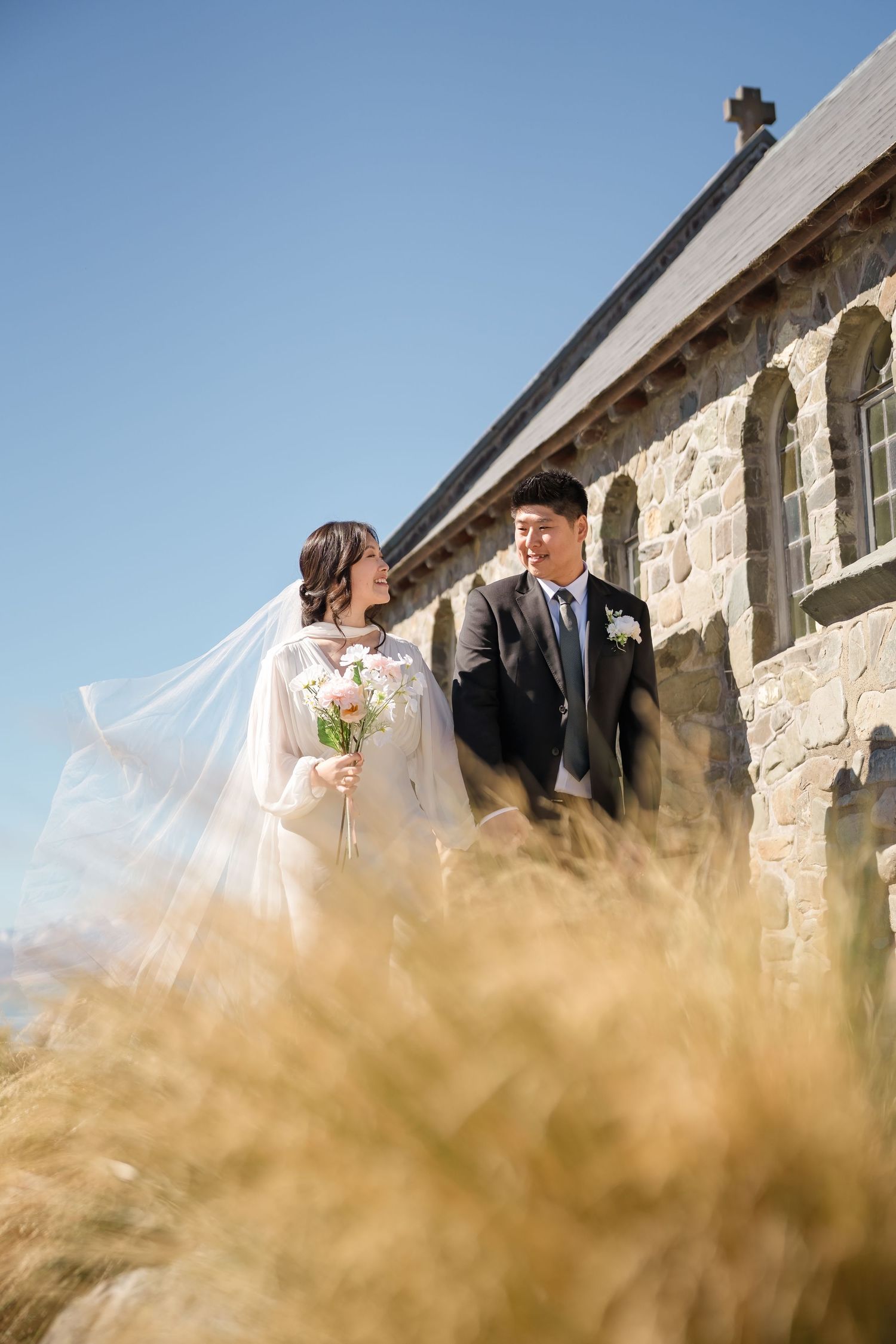 Bride and groom share a romantic moment outside a stone church, her veil flowing in the breeze under a clear blue sky.