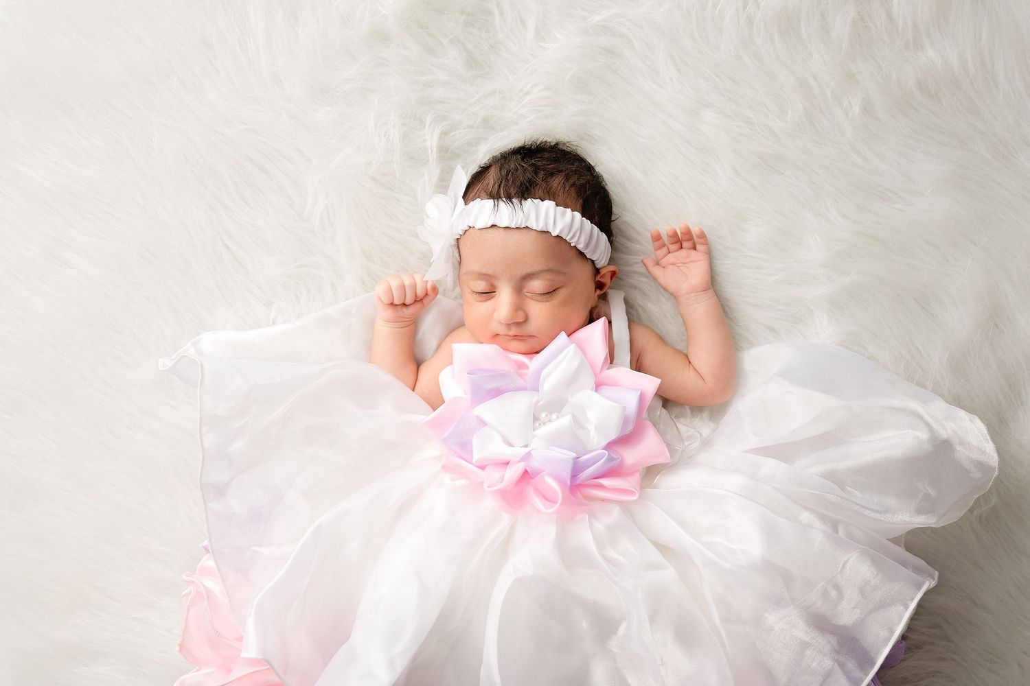 Sleeping newborn baby girl in white tutu dress and pink bow headband on fluffy white background.